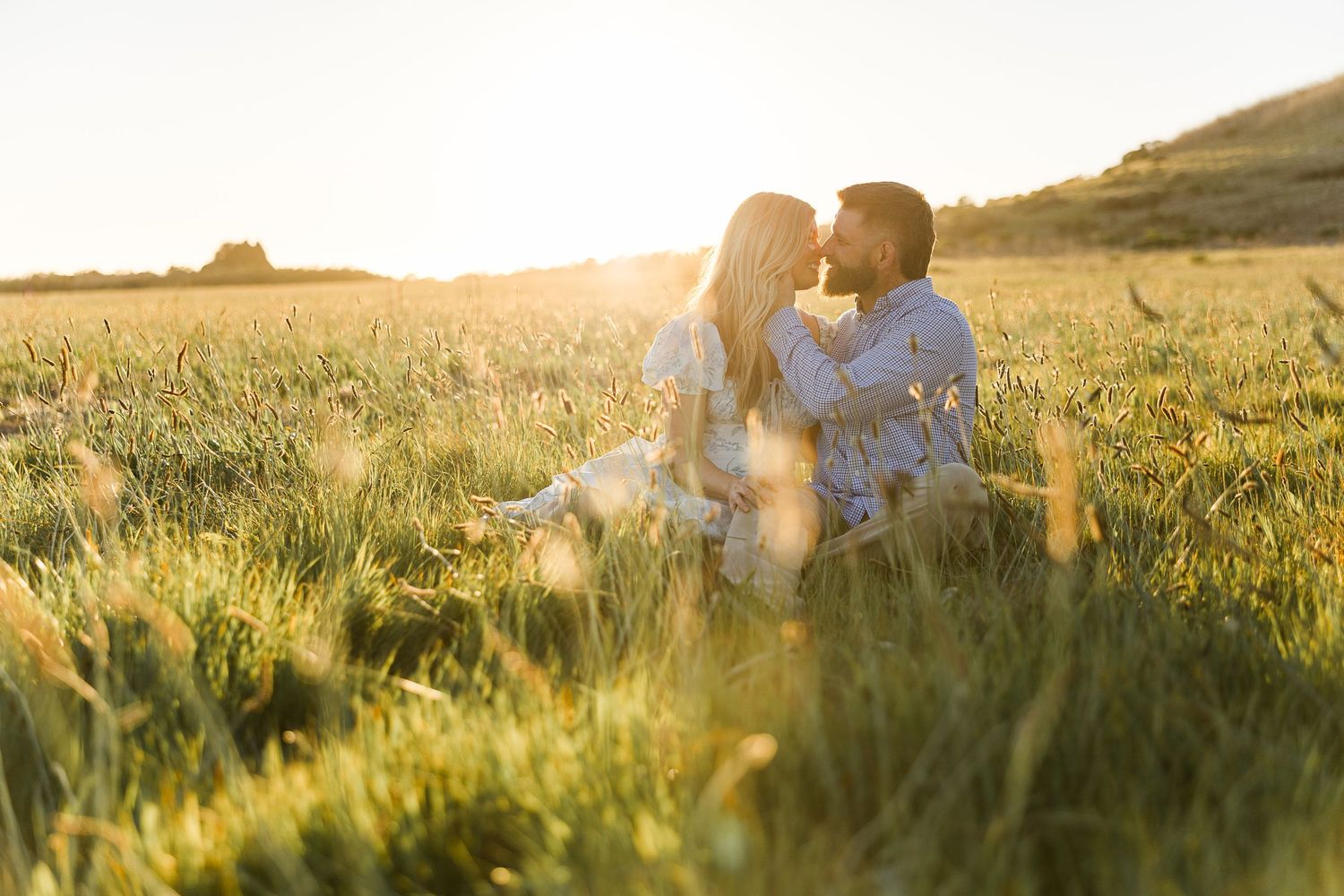 A couple sits together in a sunlit meadow during golden hour as sunlight streams through tall grass.
