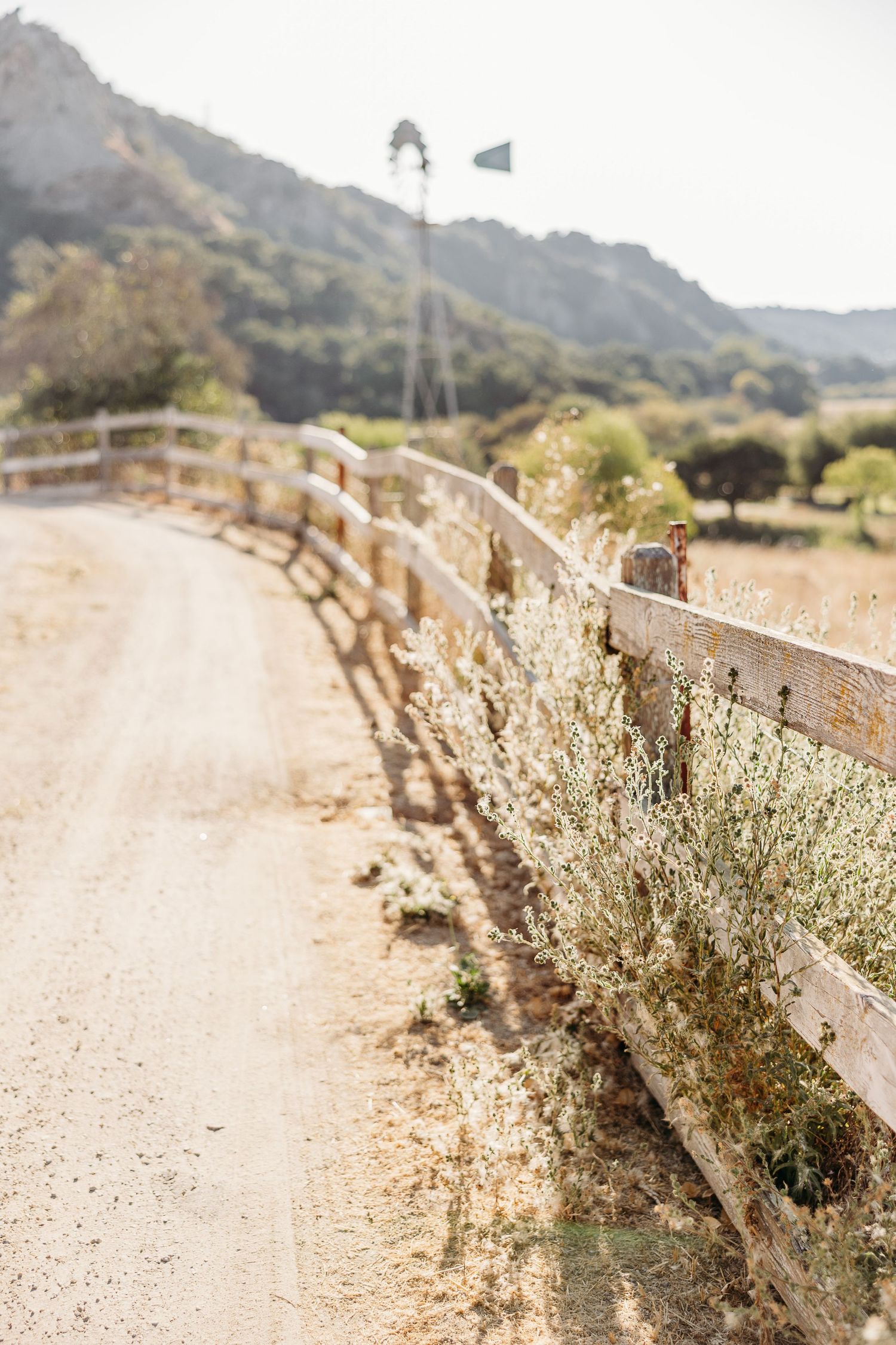 Rustic wooden fence along dirt path with windmill and mountain vista in background.