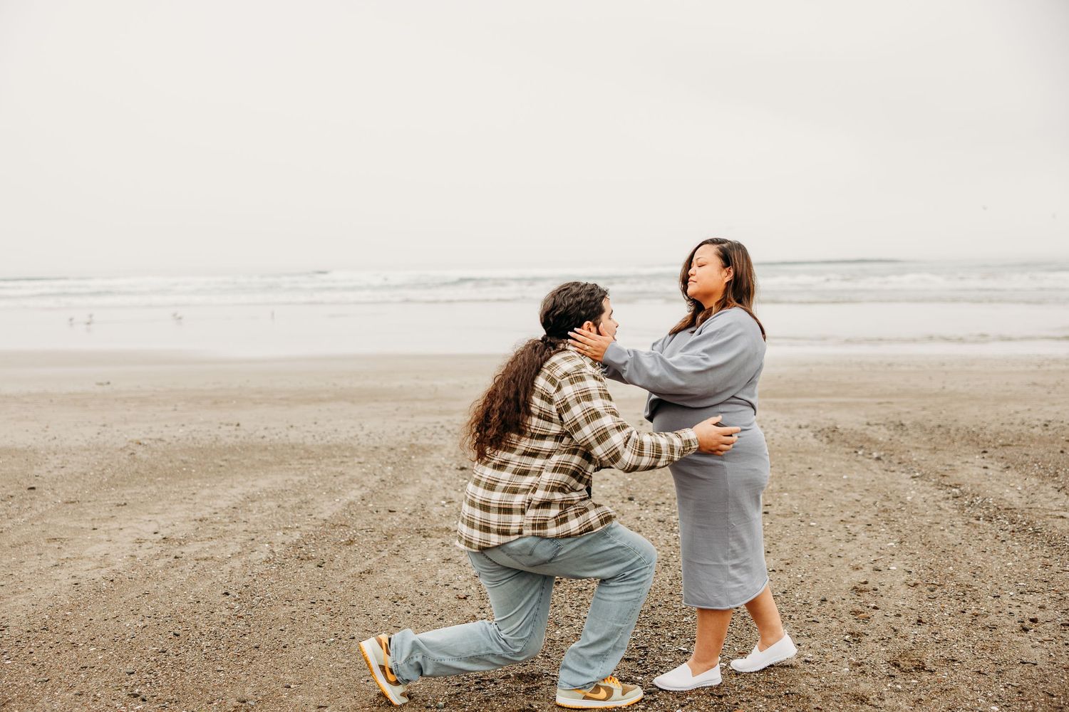 A couple shares a romantic moment on a misty beach as one partner proposes while kneeling in the sand.
