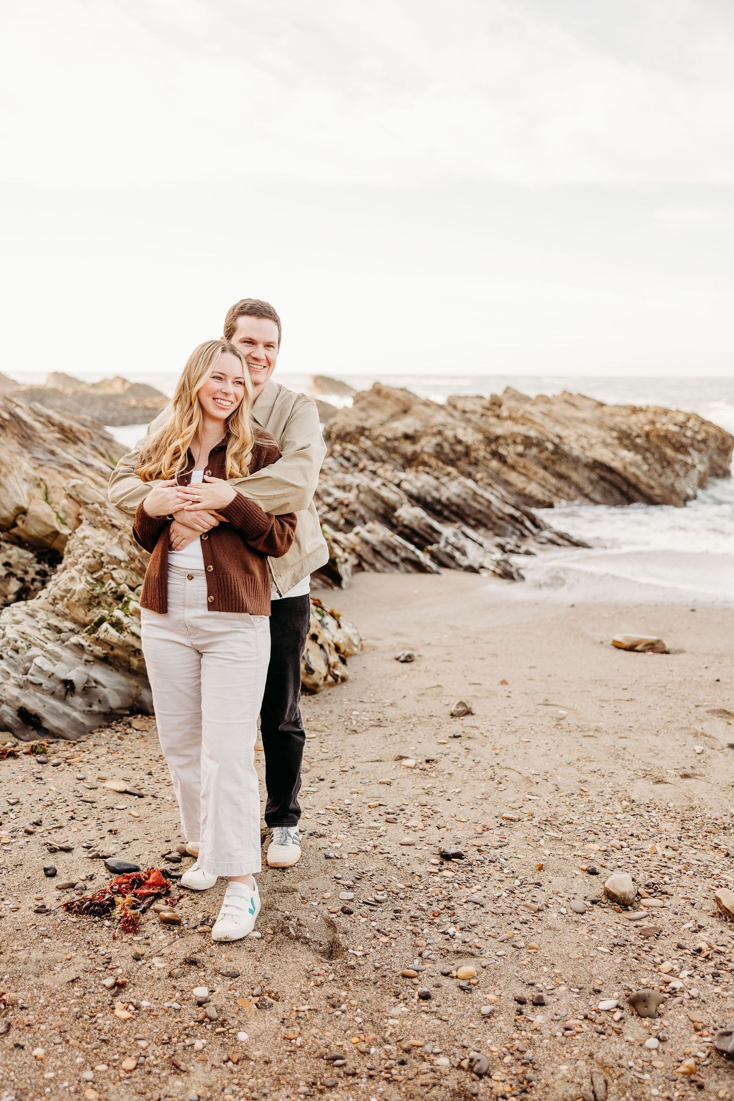 Couple walking together on a rocky beach during sunset with dramatic coastal cliffs in the background.