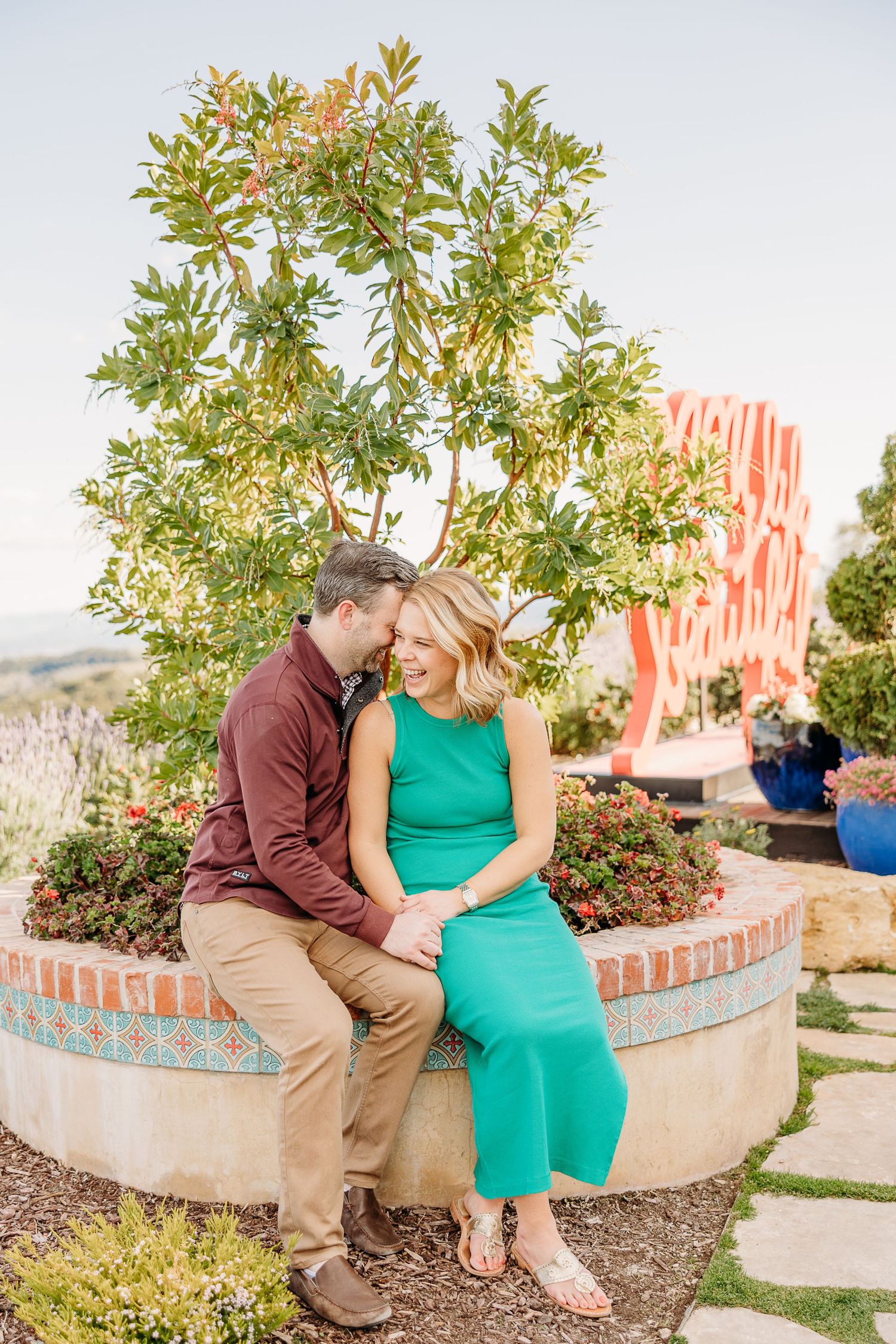 A couple shares an intimate moment while sitting on a brick planter with greenery and scenic views in the background.