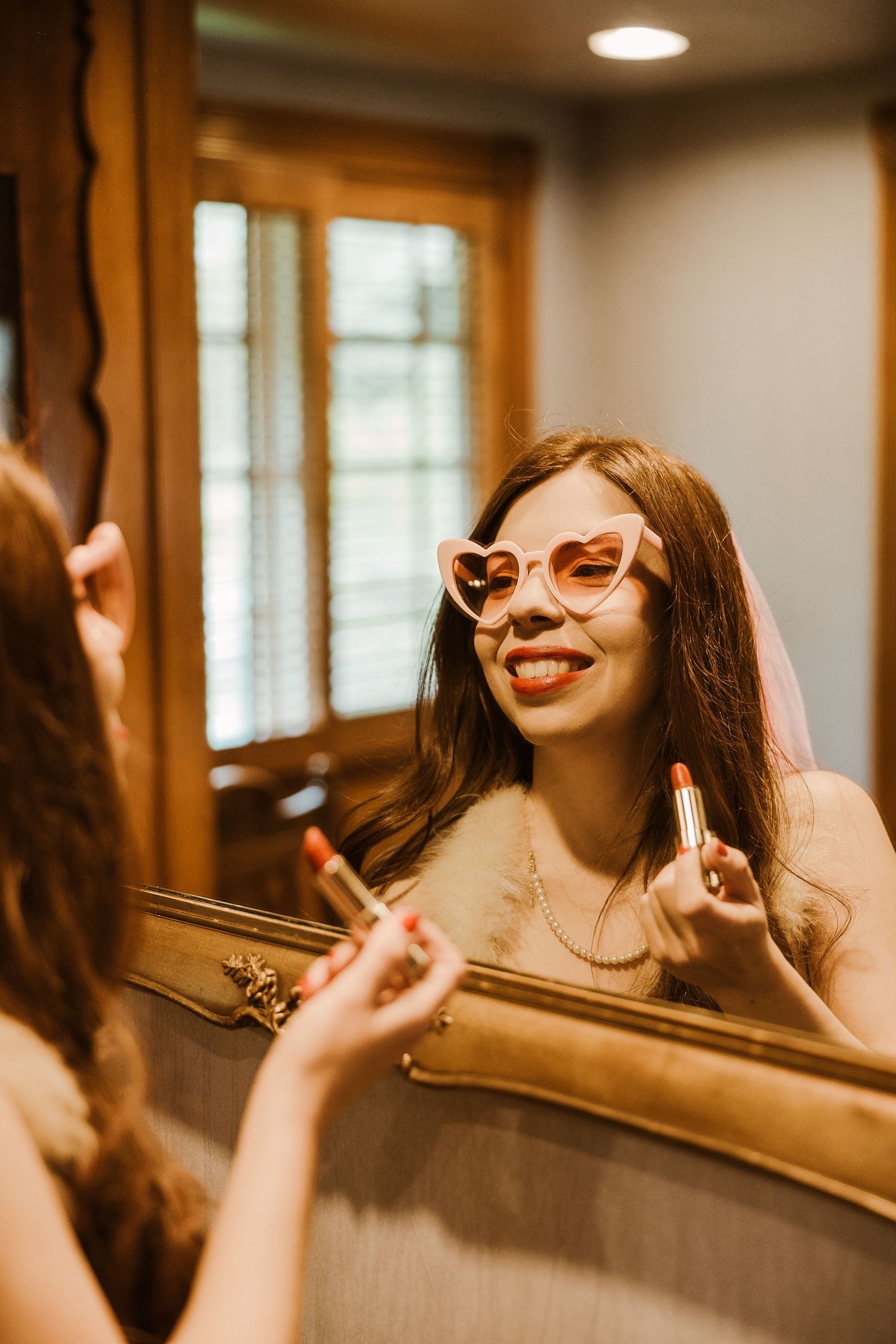 A reflection in an ornate gold mirror shows someone wearing retro cat-eye sunglasses applying lipstick in warm lighting.