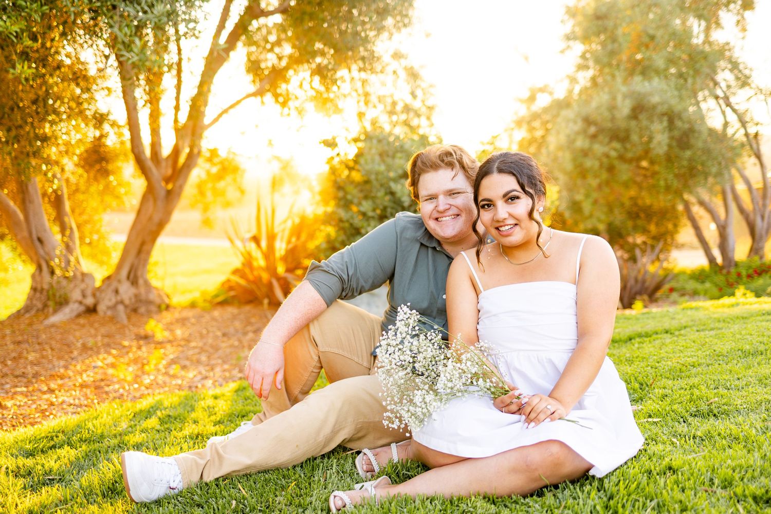 Couple sitting together on grass during golden hour sunset with flowers and trees in background.