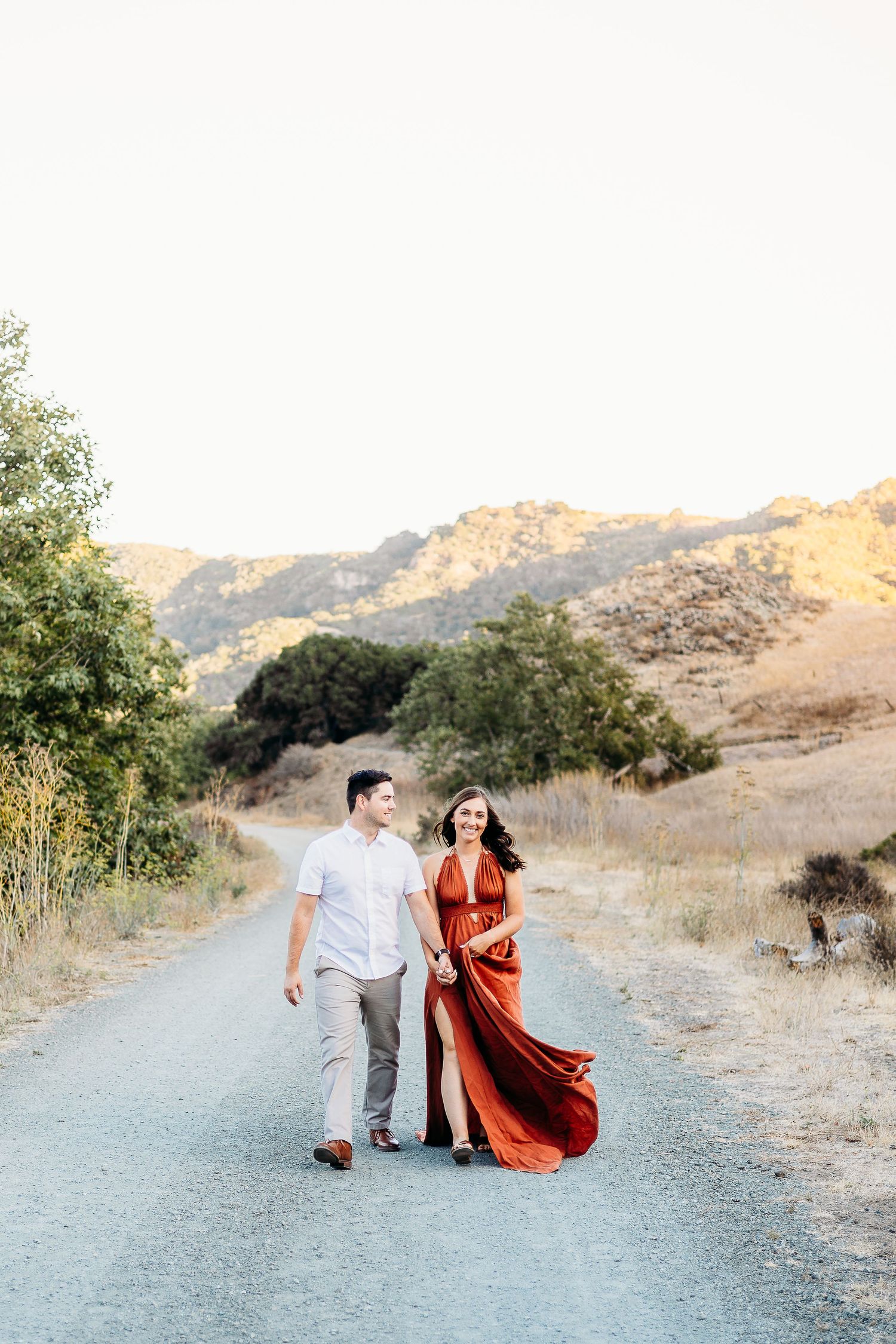 Couple walks along mountain path in elegant white shirt and flowing rust dress.
