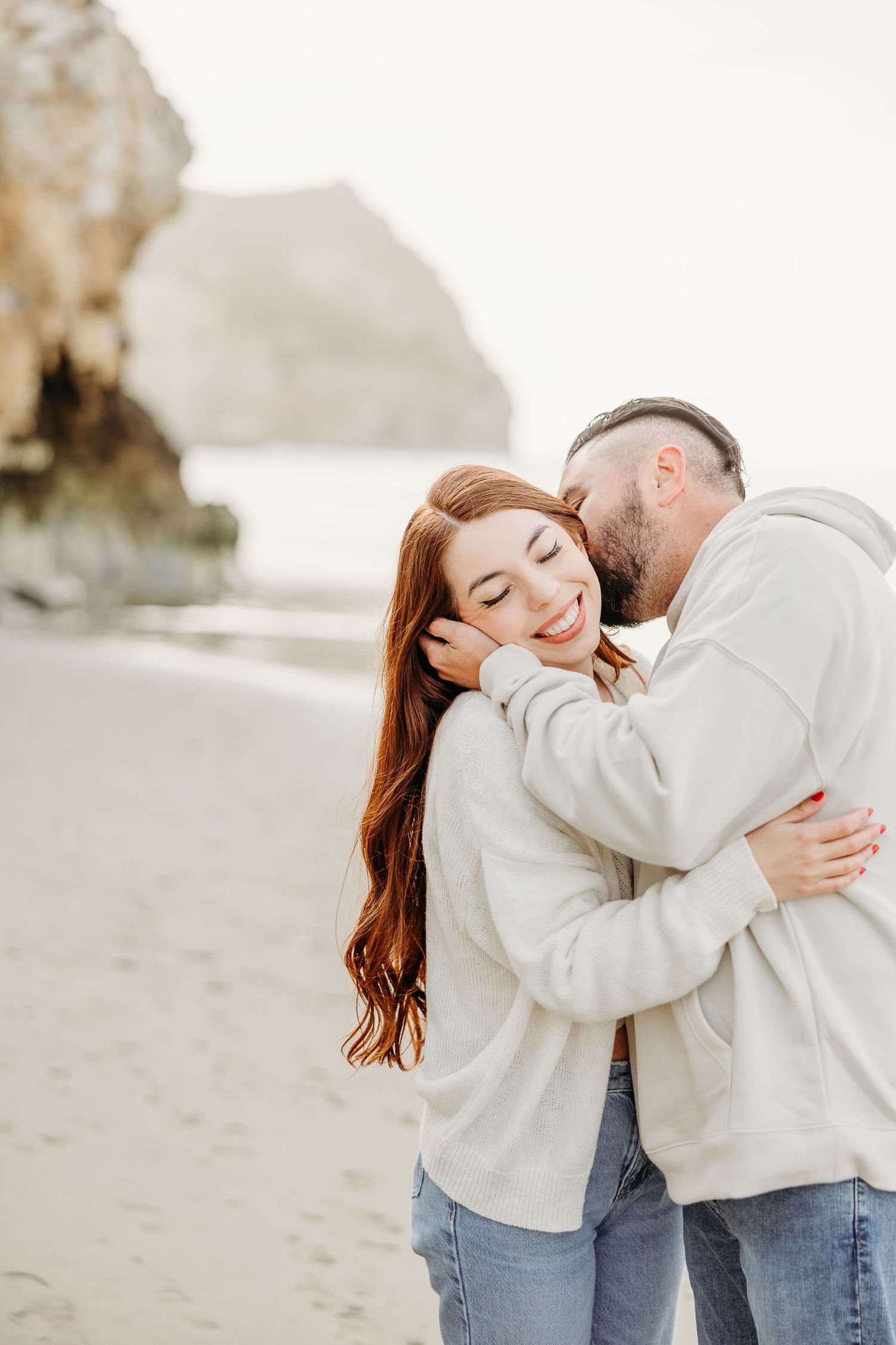 Couple shares tender moment together on beach with coastal rocks in background.