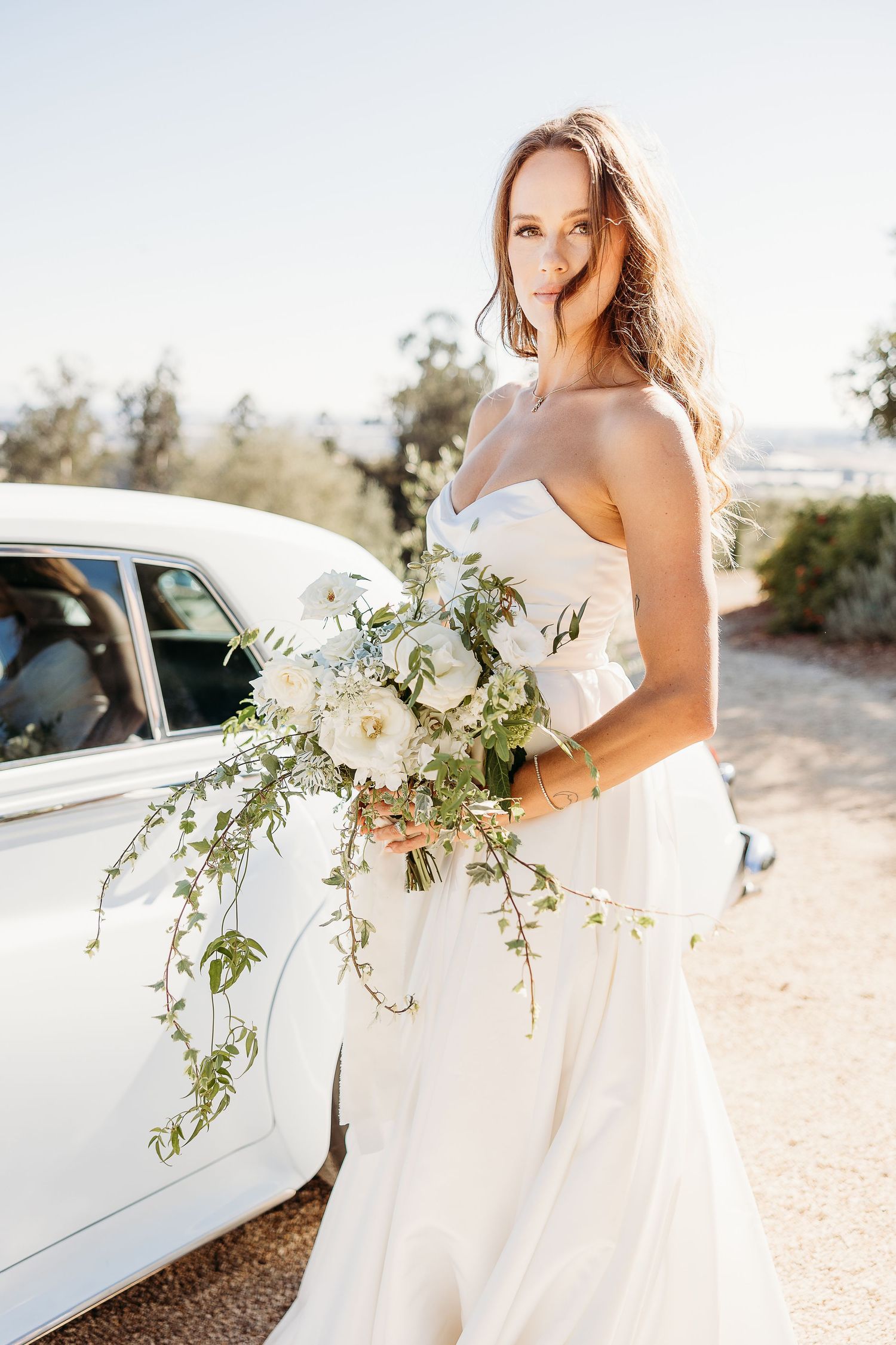 Bride in white strapless gown holds cascading bouquet next to vintage car.