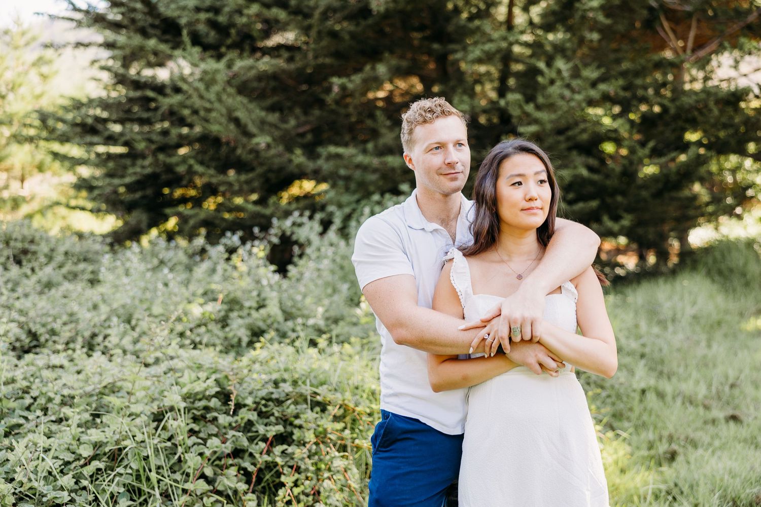 A couple poses together in front of green shrubbery and trees during a sunny outdoor photoshoot.