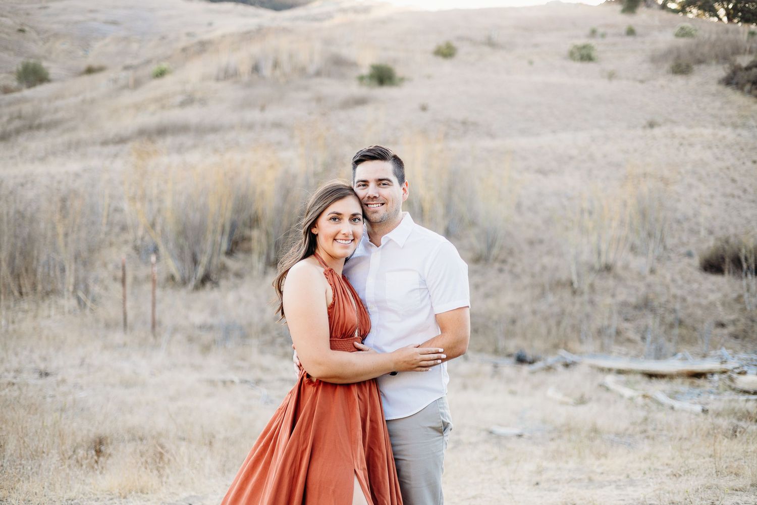 A couple embraces on a rustic trail with golden hills in the background.