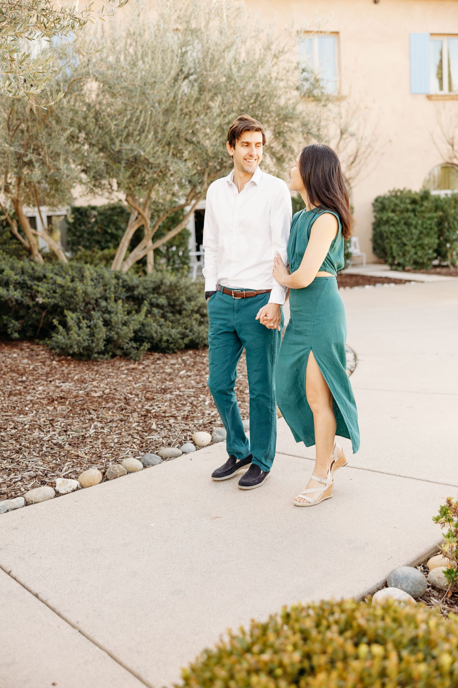 A couple in coordinated outfits standing on a pathway with Mediterranean landscaping.