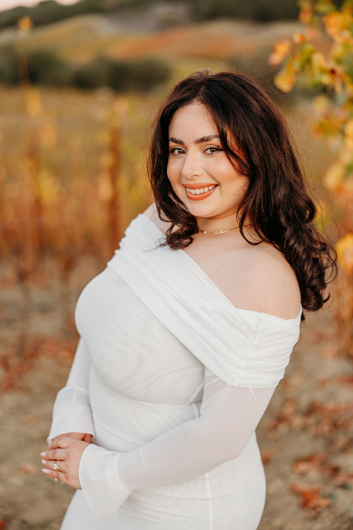 A woman in a flowing white dress poses gracefully against an autumn backdrop during golden hour.