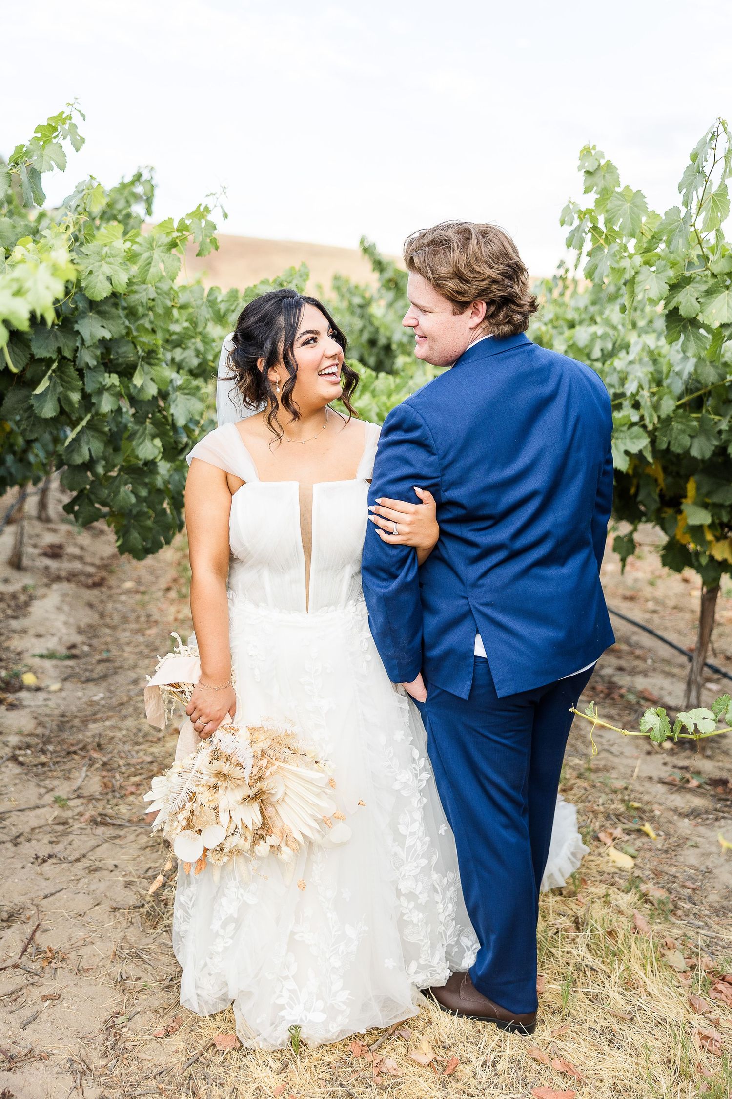 Joyful couple share loving moment in vineyard with bride holding dried floral bouquet.