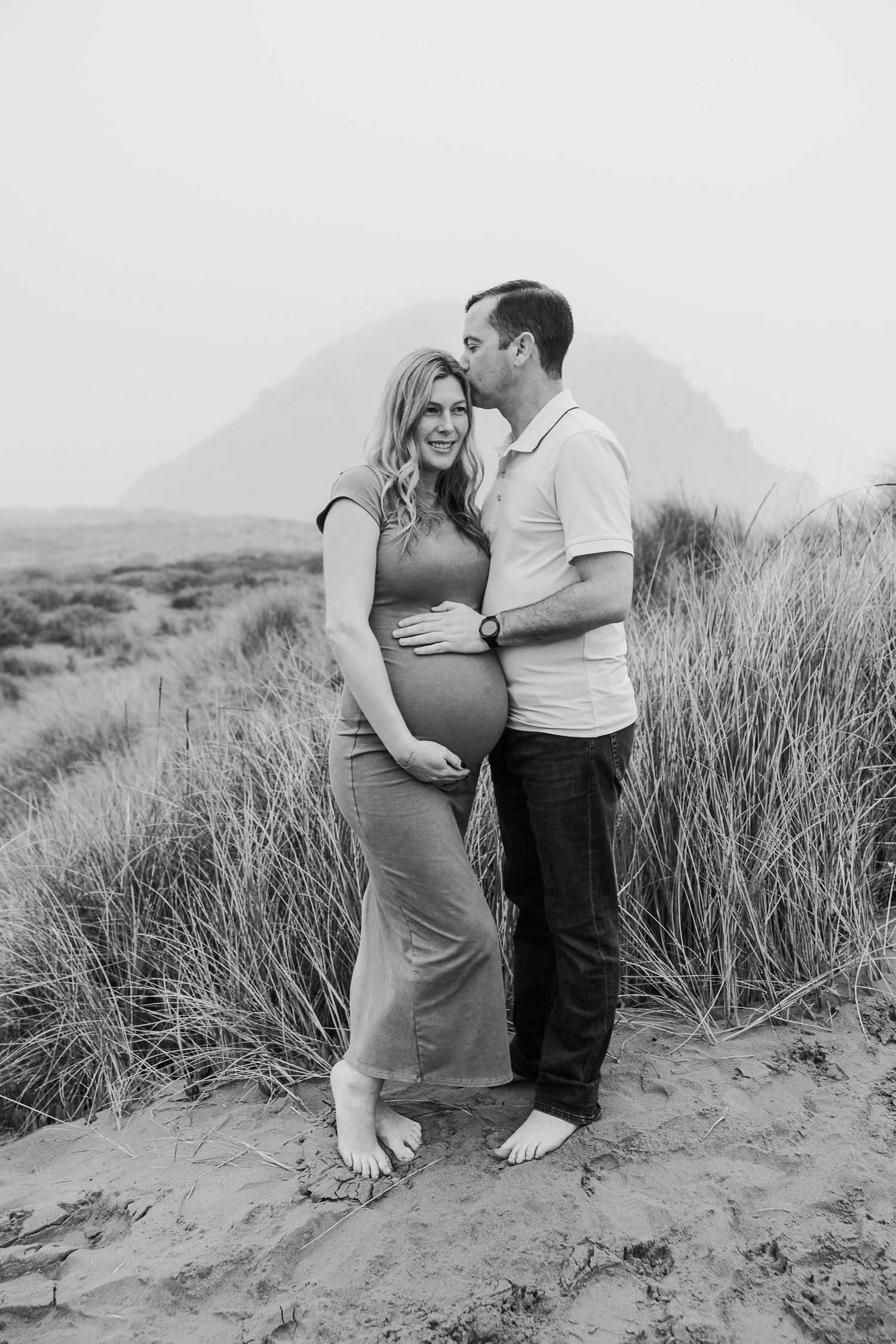 Full-length maternity portrait of couple standing among beach grasses.