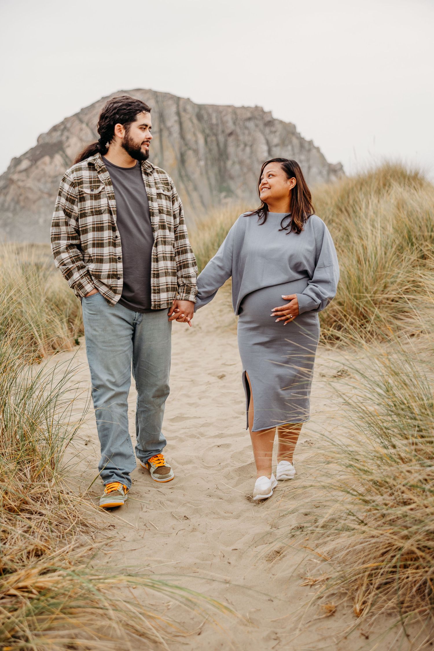 A couple walking on a sand path through beach grass with a mountain backdrop on an overcast day.