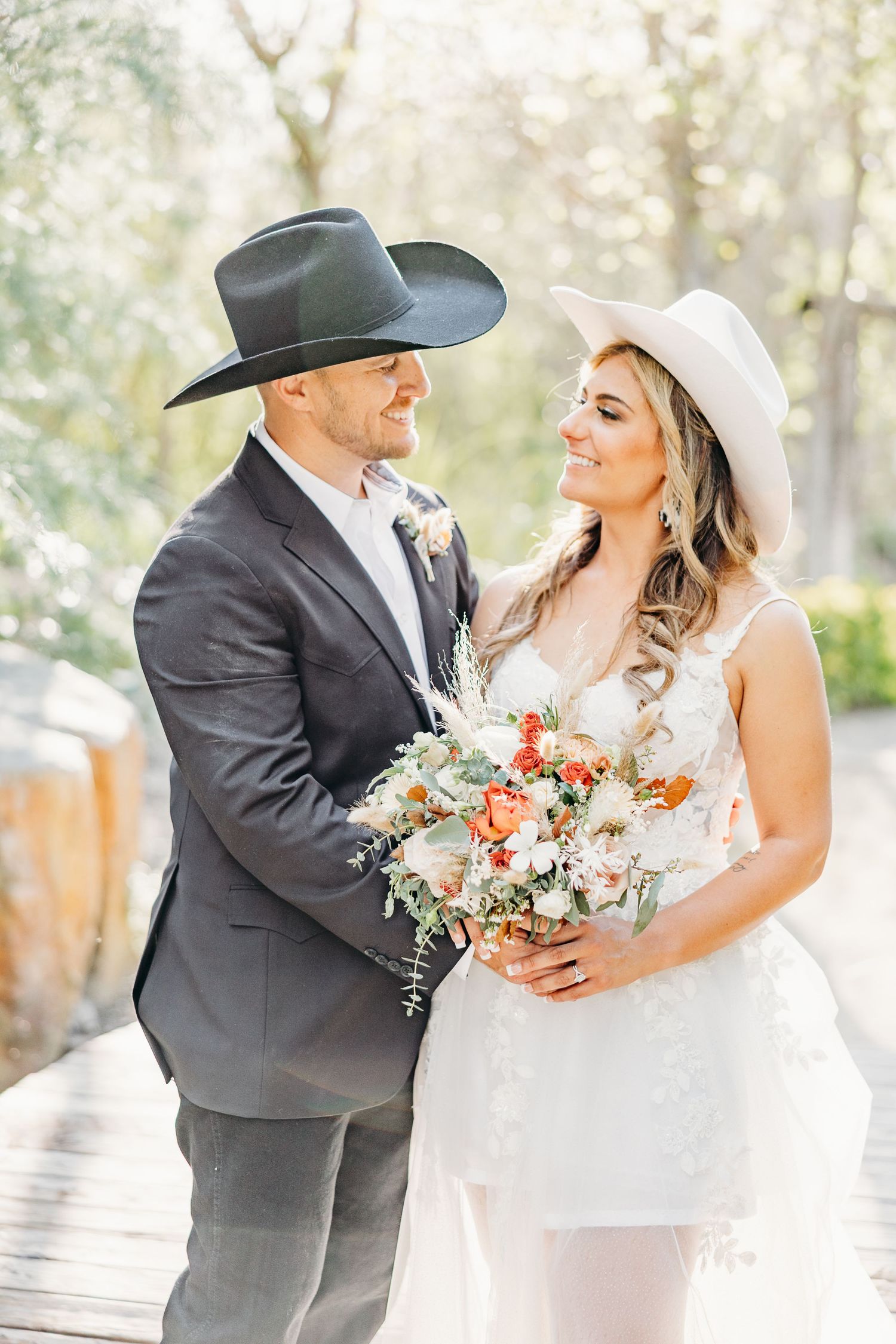 Newlyweds in cowboy hats share an intimate moment while holding a floral bouquet on a wooden path.
