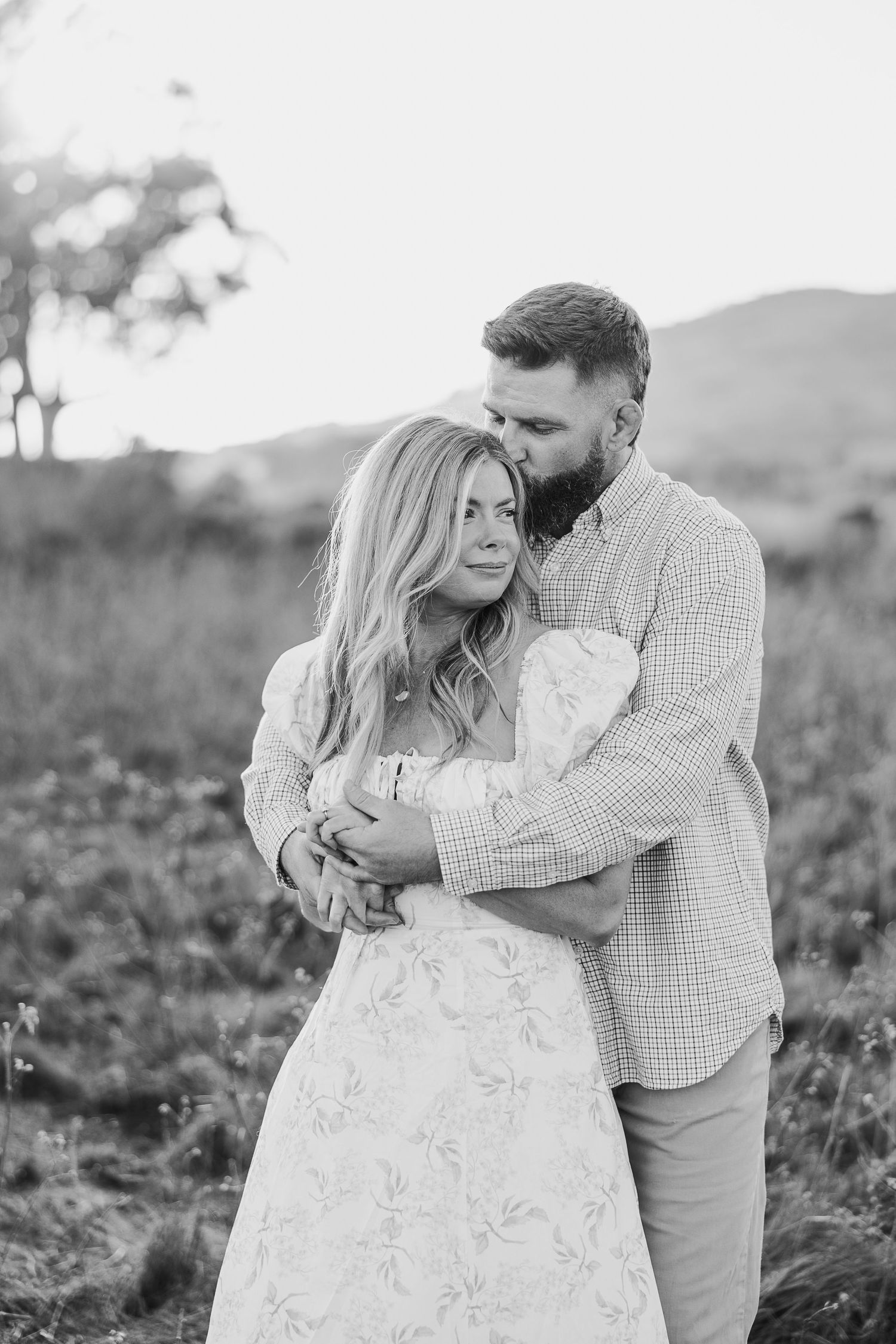 A romantic black and white portrait of a couple embracing in a field with mountains in the background.