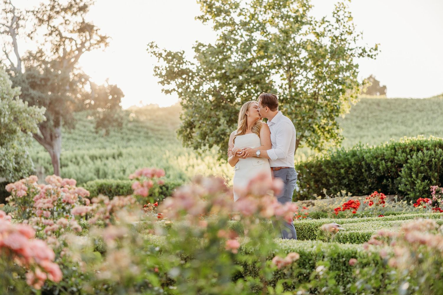 Two people share an intimate moment among blooming roses in a lush garden setting at sunset.