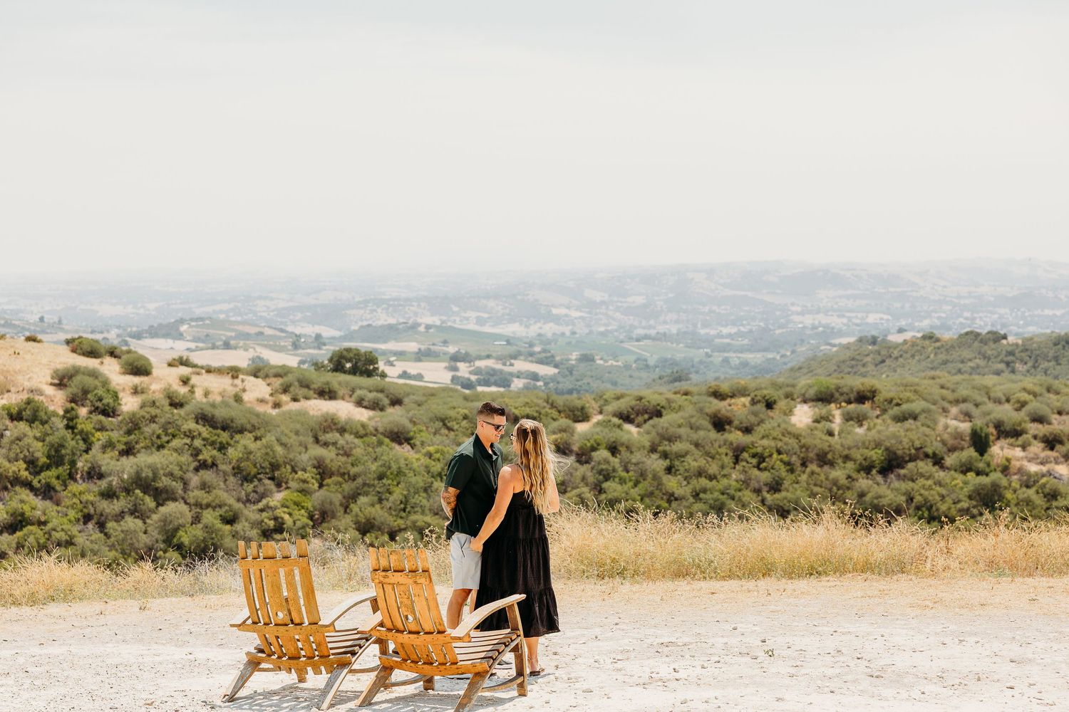A scenic panoramic view of rolling hills and mountains with a person sitting in an Adirondack chair at sunset.
