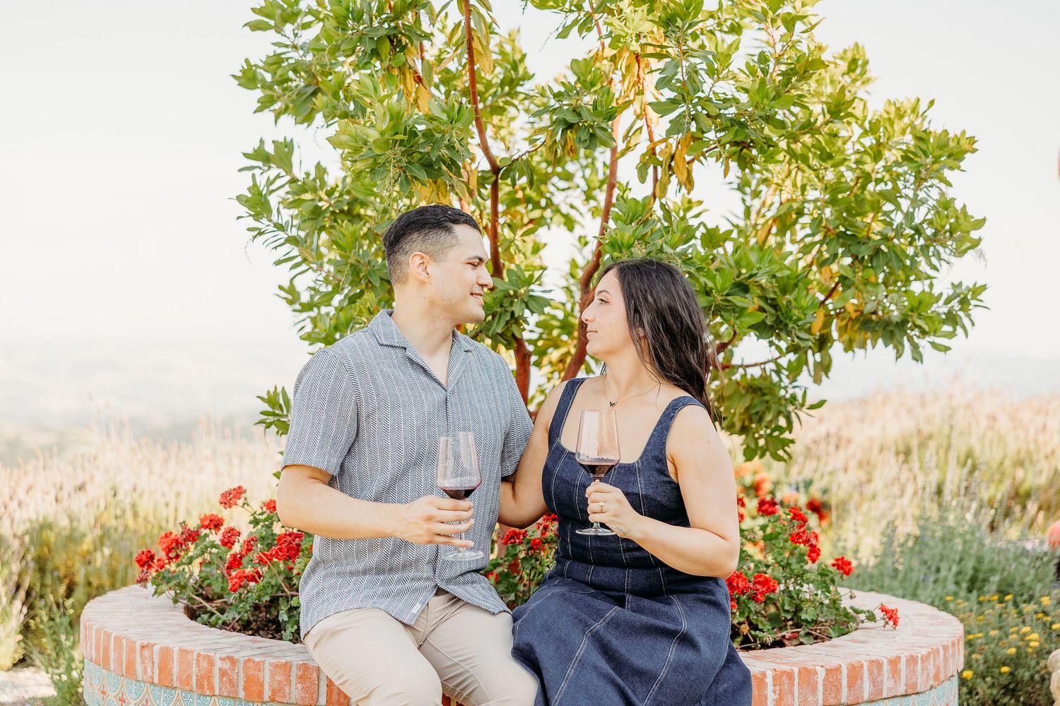 Couple sharing drinks and laughing together on outdoor patio with flowering plants.