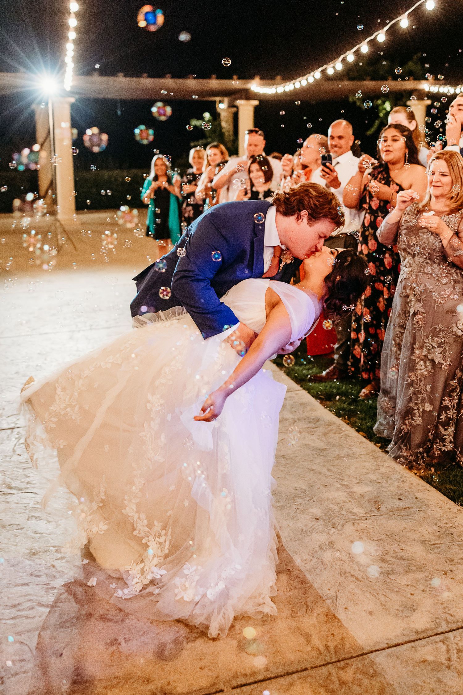 Romantic dip kiss during first dance with bubbles floating around at outdoor wedding reception.