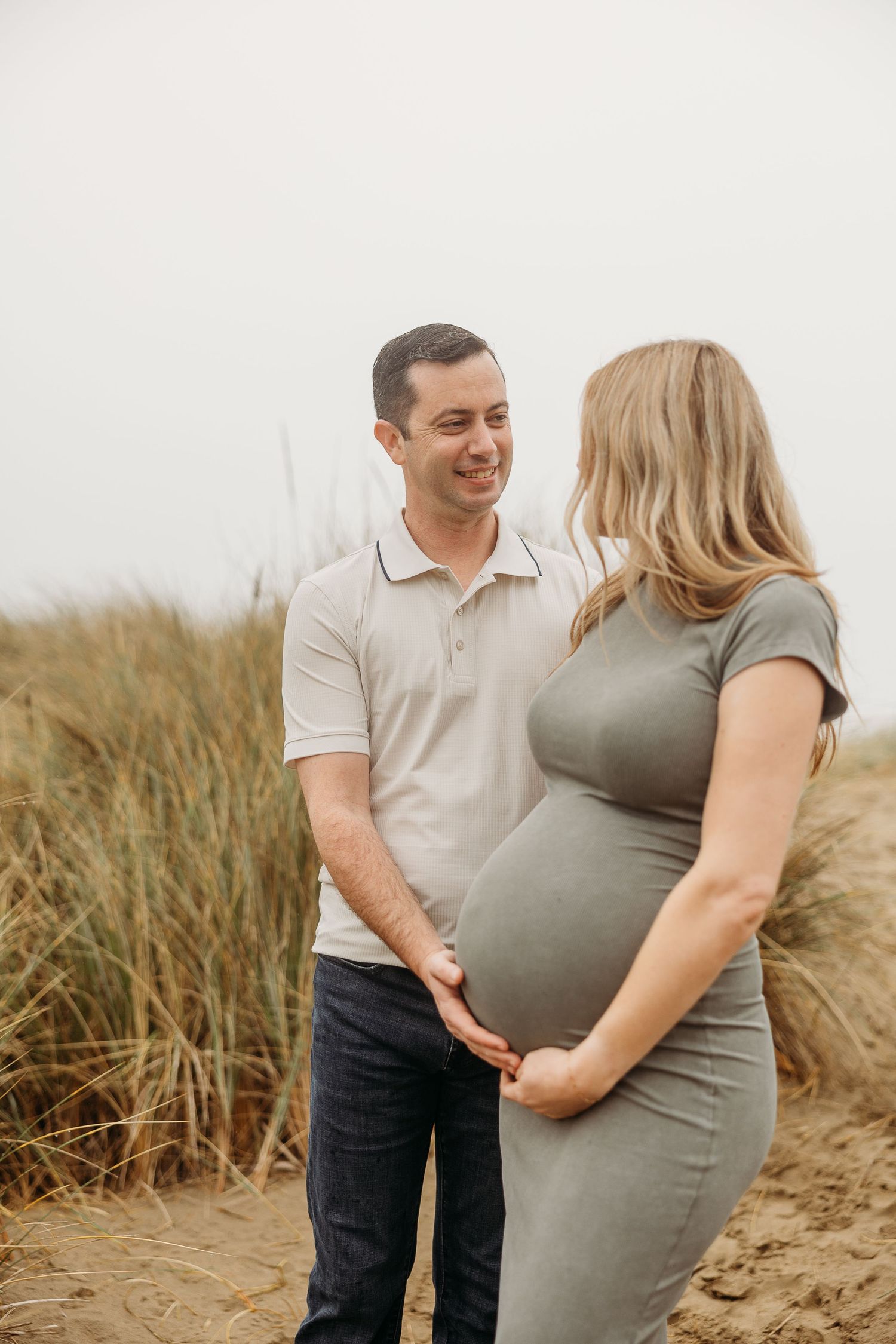 A couple stands together on sandy dunes during their maternity photoshoot.