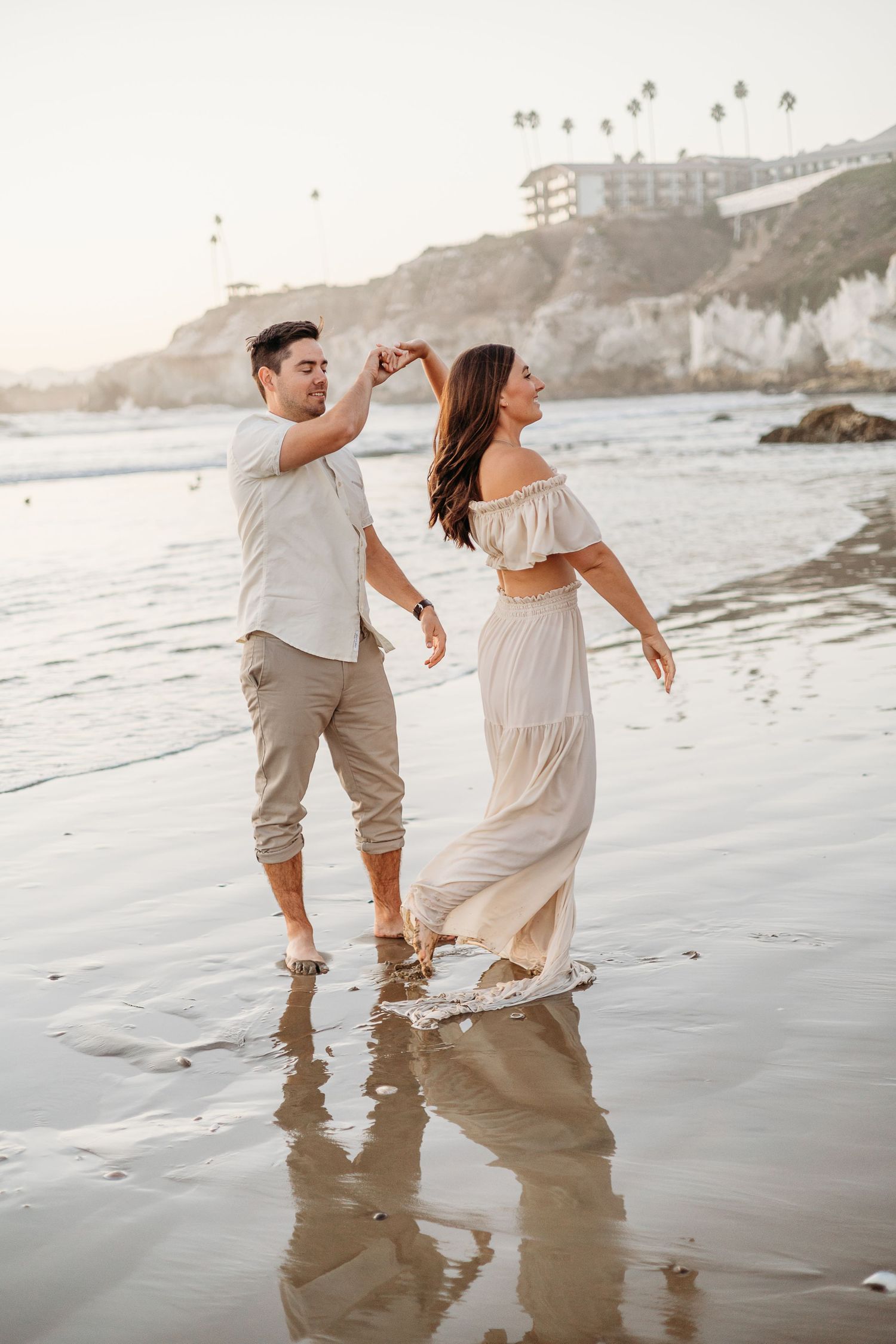 A romantic couple dancing barefoot on a beach at sunset with white cliffs and buildings in the background.