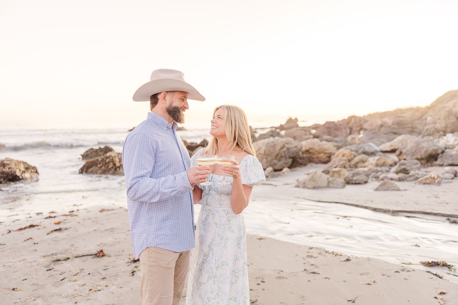 A romantic couple shares a tender moment on a rocky beach at sunset wearing light summer attire.