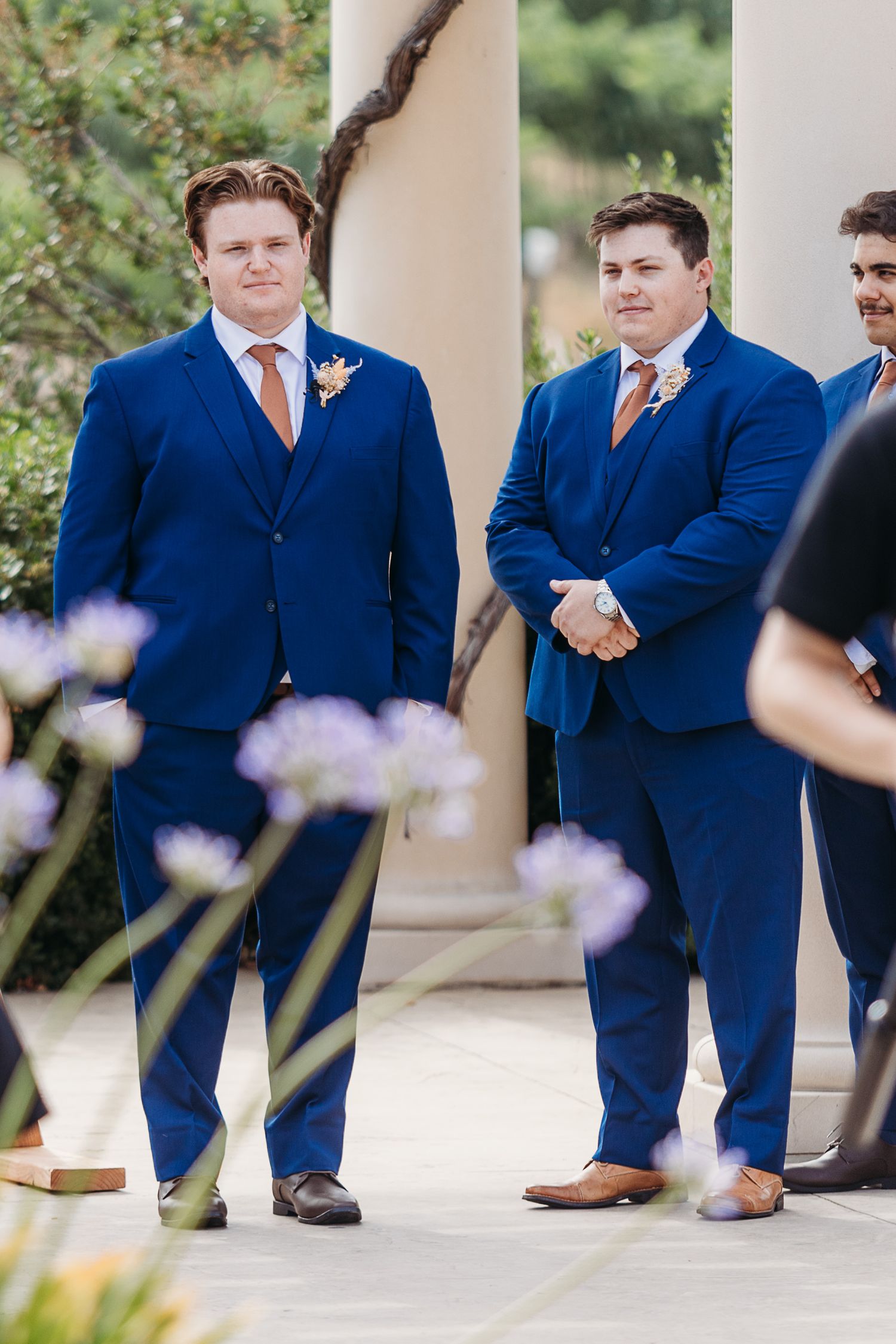 Groomsmen in navy suits with orange ties stand under outdoor pergola.