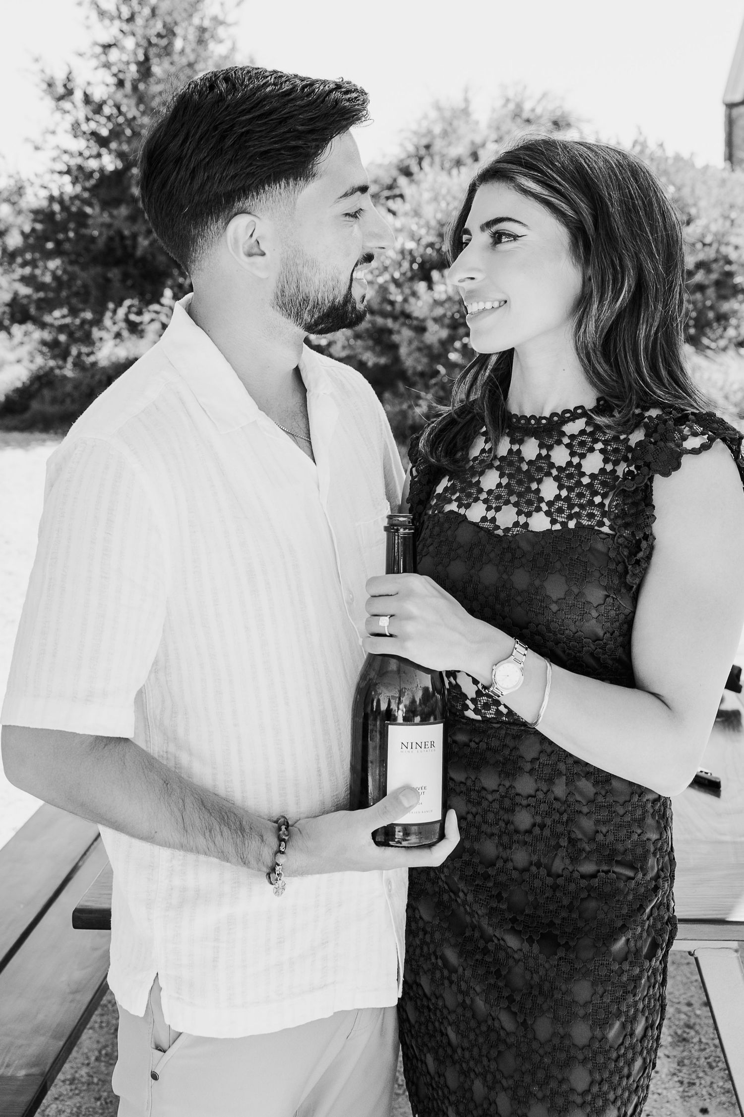A romantic black and white photo of a couple sharing a moment while holding a wine bottle outdoors.