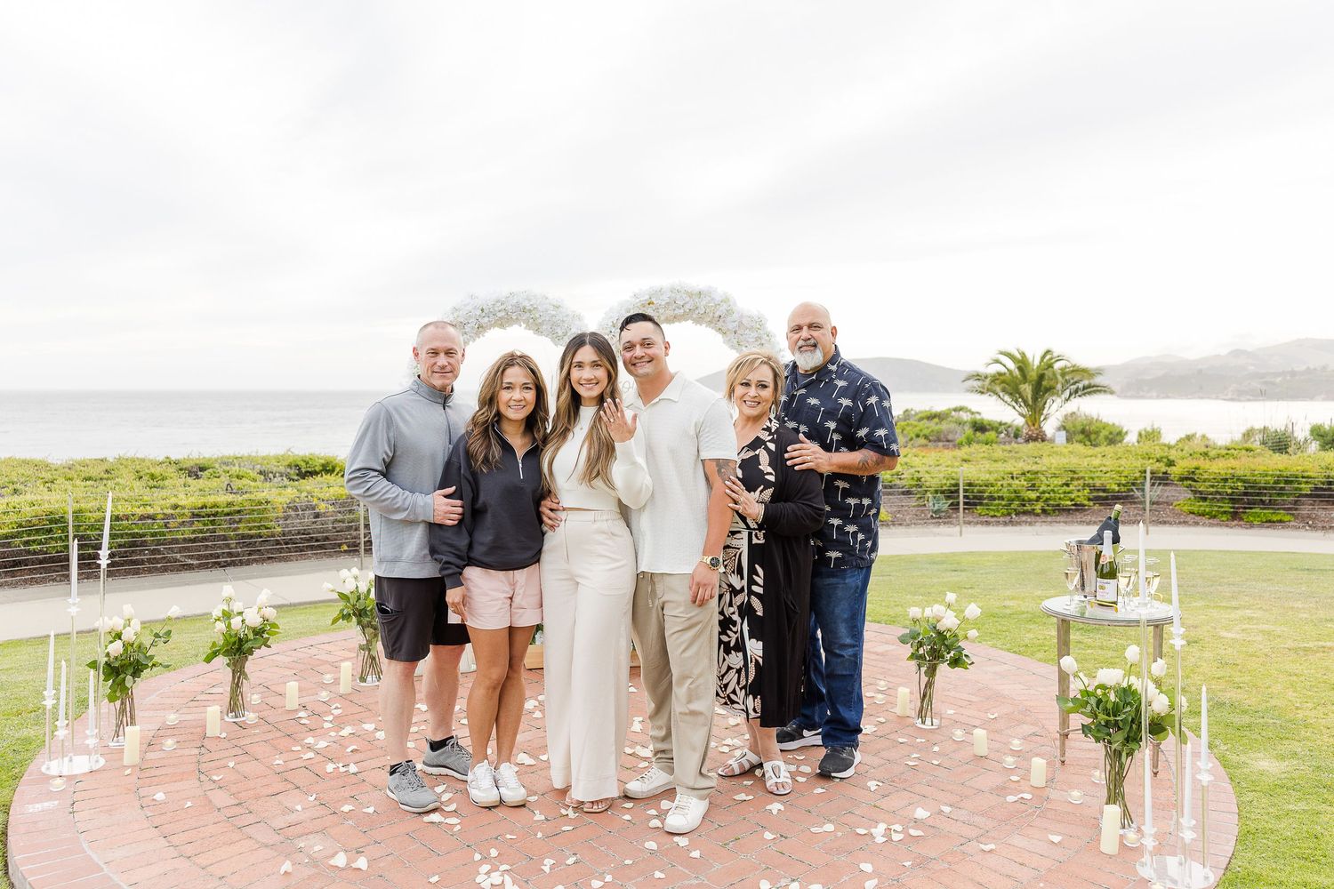 A group of people gather for photos on a brick patio with coastal scenery in the background.