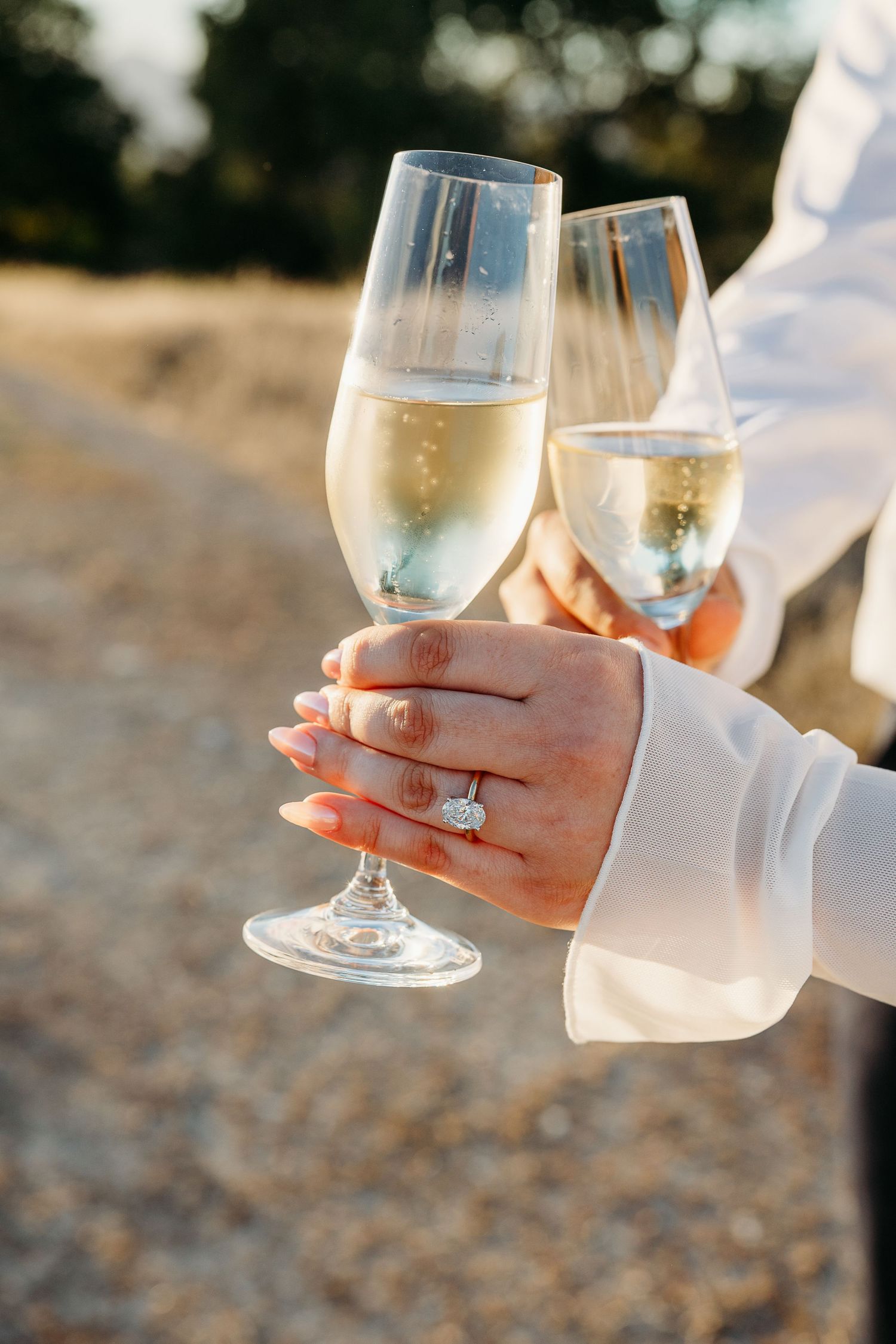 Two champagne flutes clink together in celebration as hands wearing an engagement ring hold the glasses.