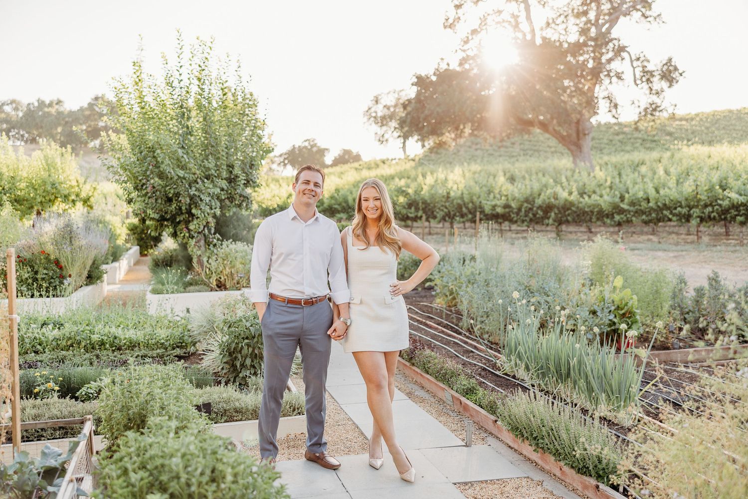 Couple walks hand in hand through a sunlit garden path surrounded by lavender and greenery at sunset.
