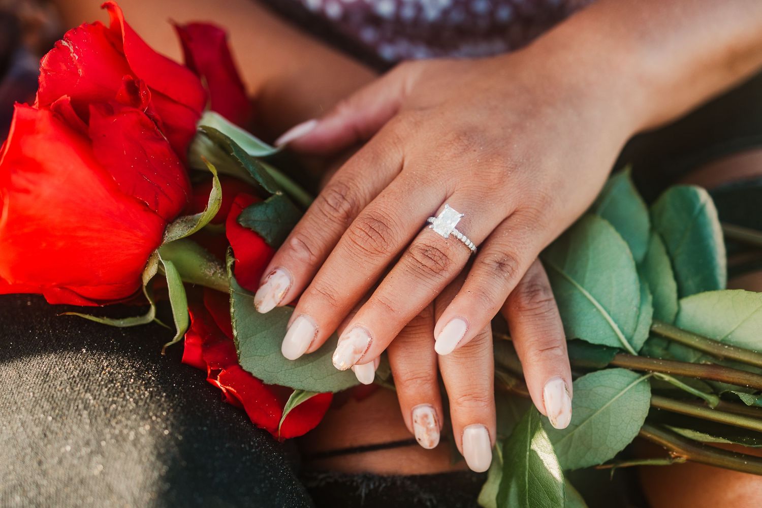 A close-up of a hand wearing an engagement ring next to red roses.