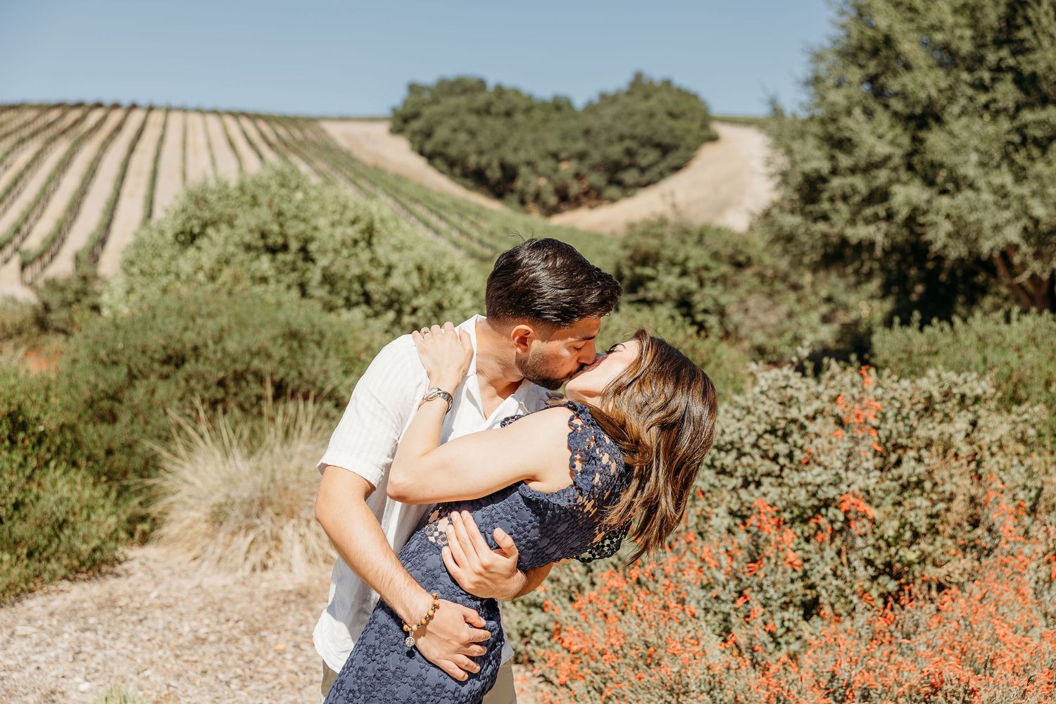 Romantic couple embracing in a sunlit vineyard with rows of grapevines and orange wildflowers.
