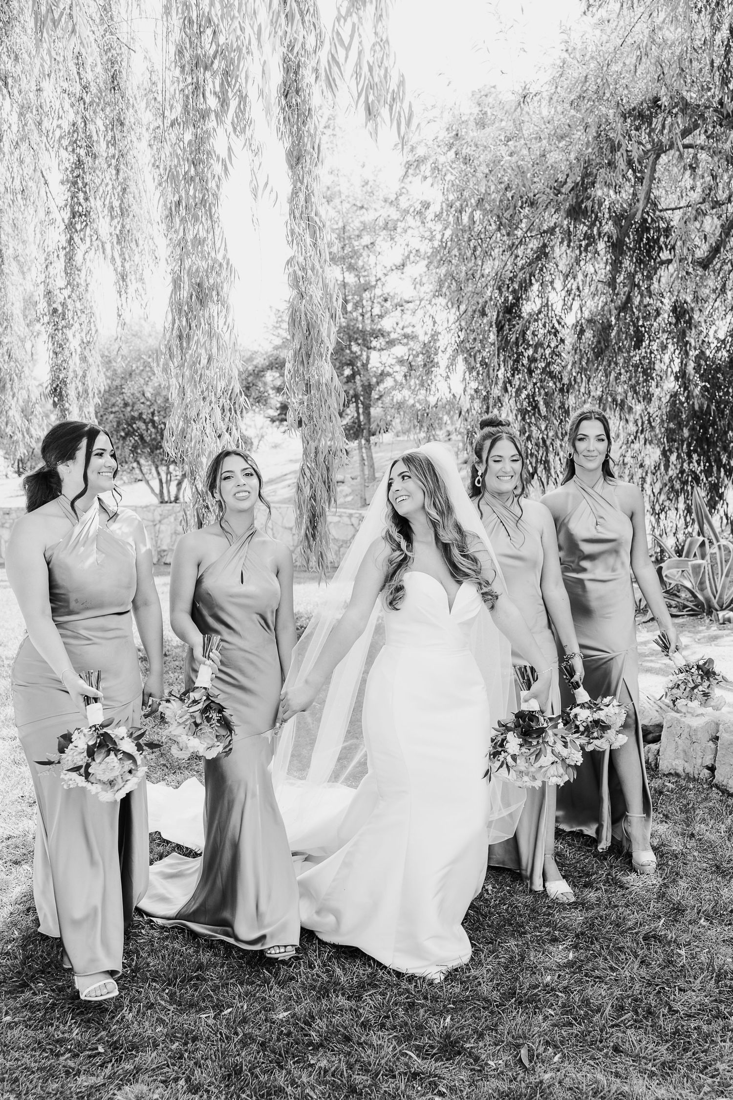 Wedding party in matching dresses poses together under trees in elegant black and white photo.