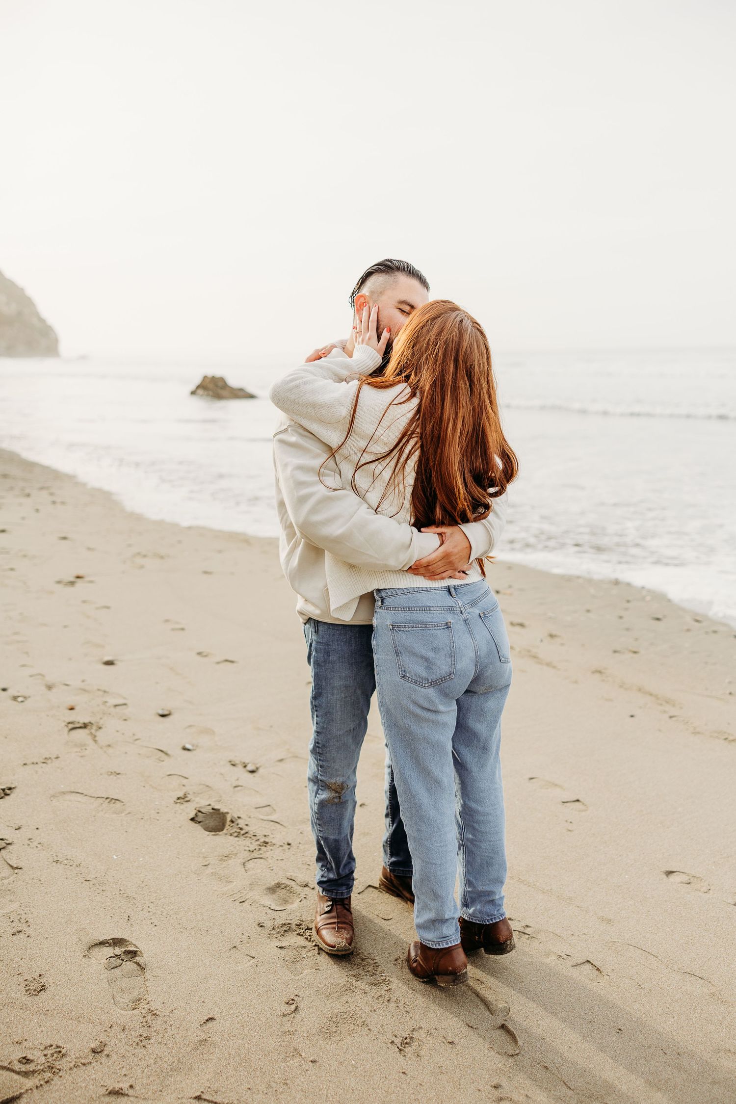 A romantic couple embraces on a misty beach wearing coordinated casual outfits with jeans and light tops.