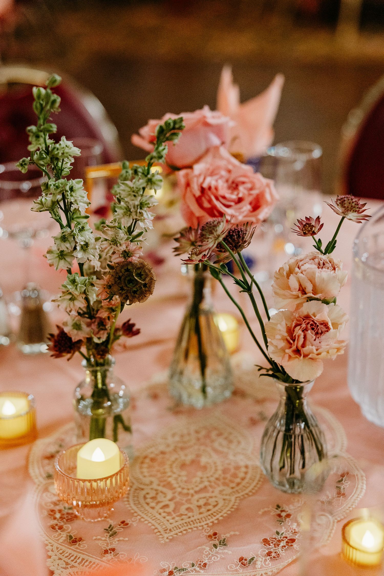 Romantic table centerpiece with pink roses and greenery in glass vases alongside glowing candles on lace tablecloth.