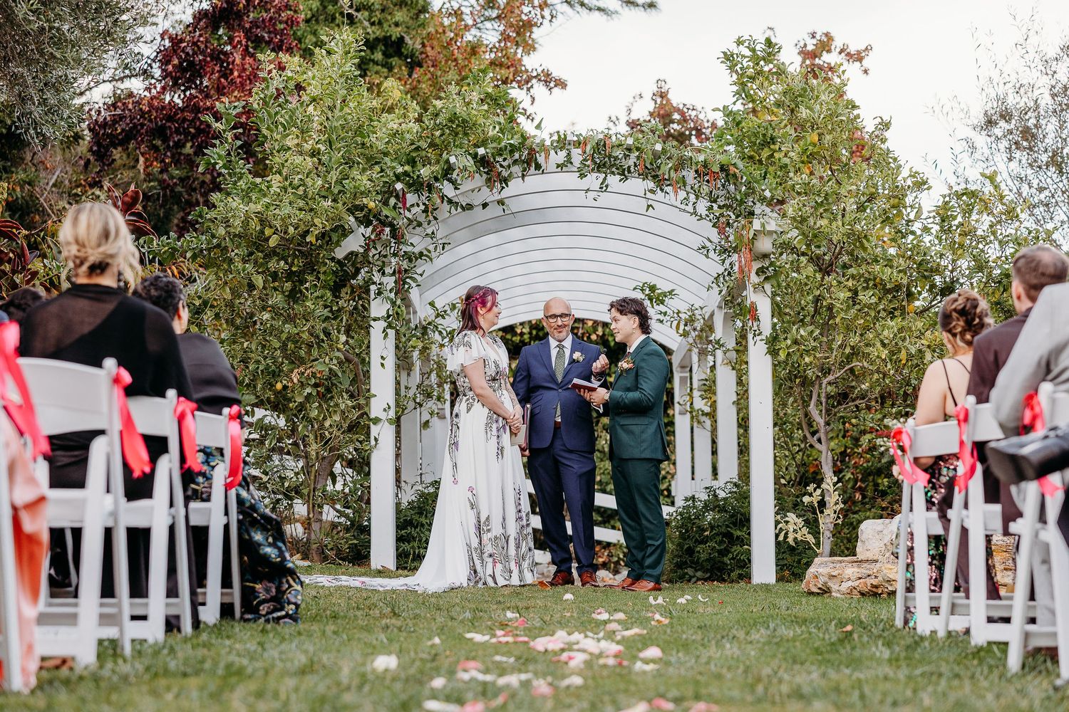 Outdoor wedding ceremony under white arch with scattered rose petals.