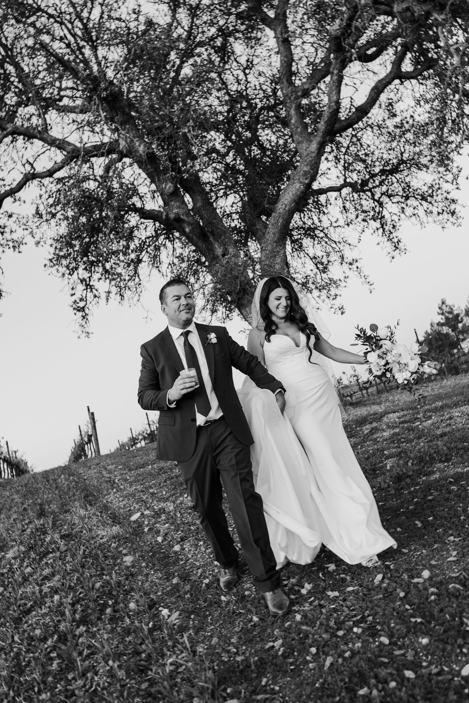 Black and white wedding photo under trees with couple in formal attire.