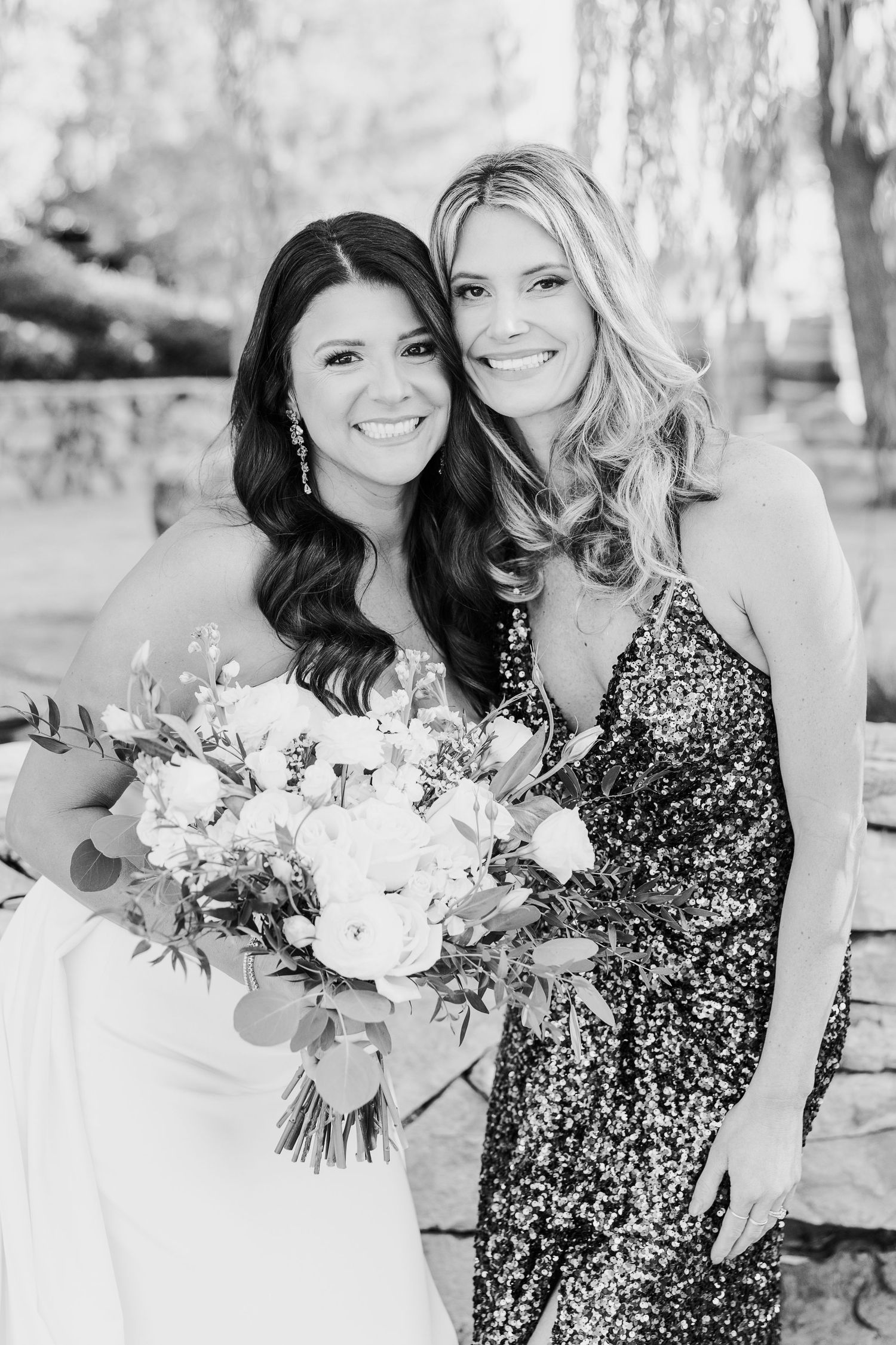 Wedding party members in white and sparkly dresses pose with floral bouquets outdoors.