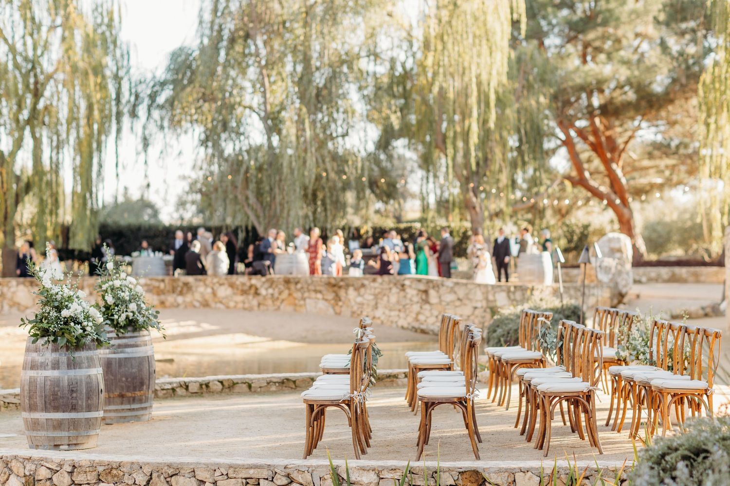 A rustic outdoor wedding ceremony setup with wooden chairs and willow trees in a garden setting.