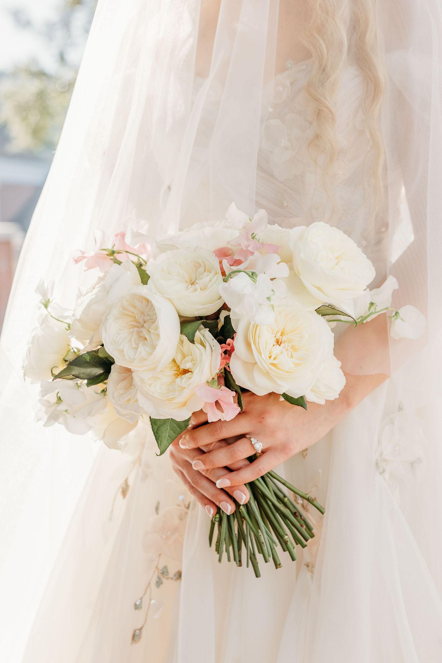 A delicate bridal bouquet of white and blush pink peonies and garden roses held against a flowing tulle wedding dress.