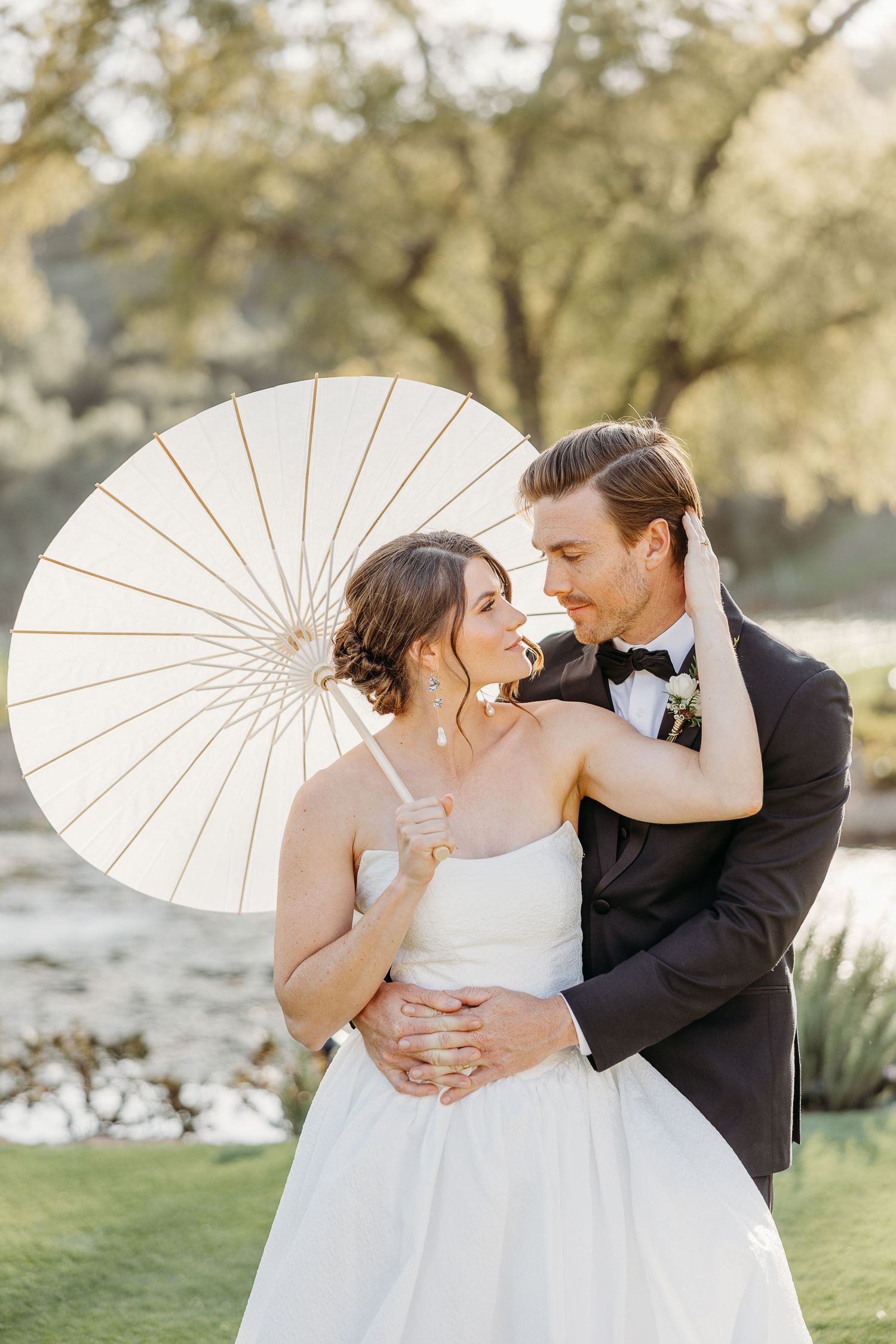 A romantic wedding couple poses outdoors with a white parasol in a sunlit garden setting.