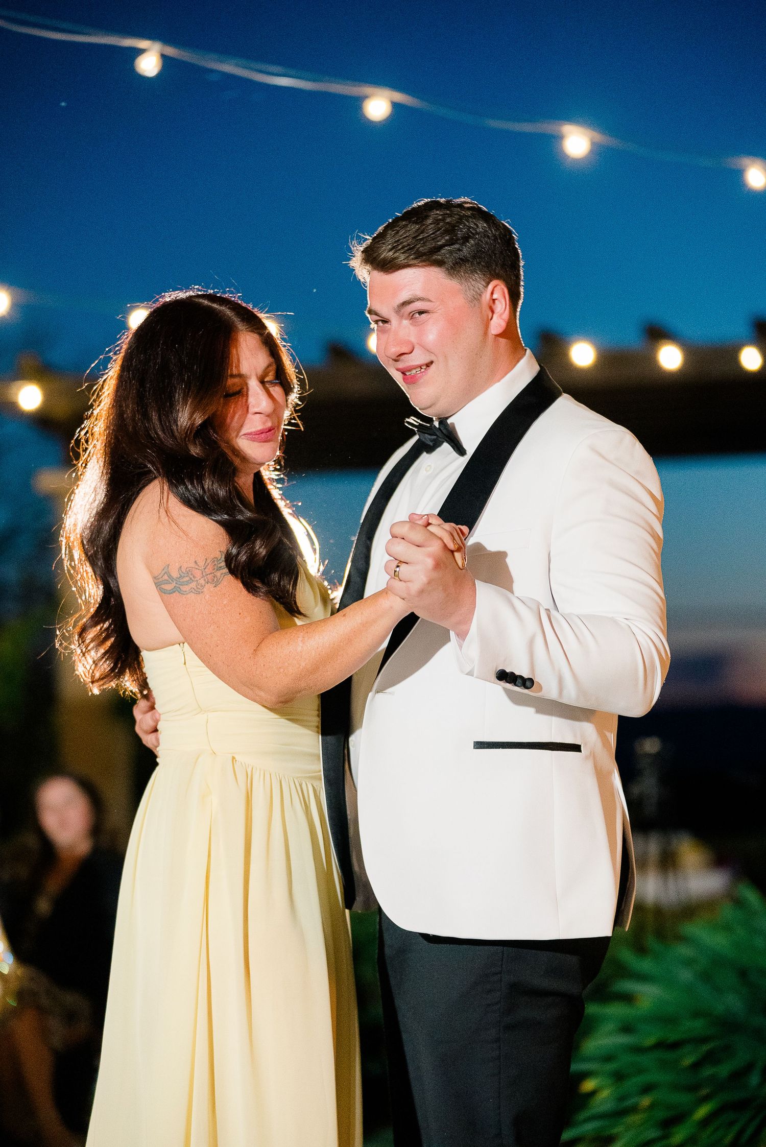 Groom in white tuxedo dances with woman in yellow dress under string lights at evening reception.