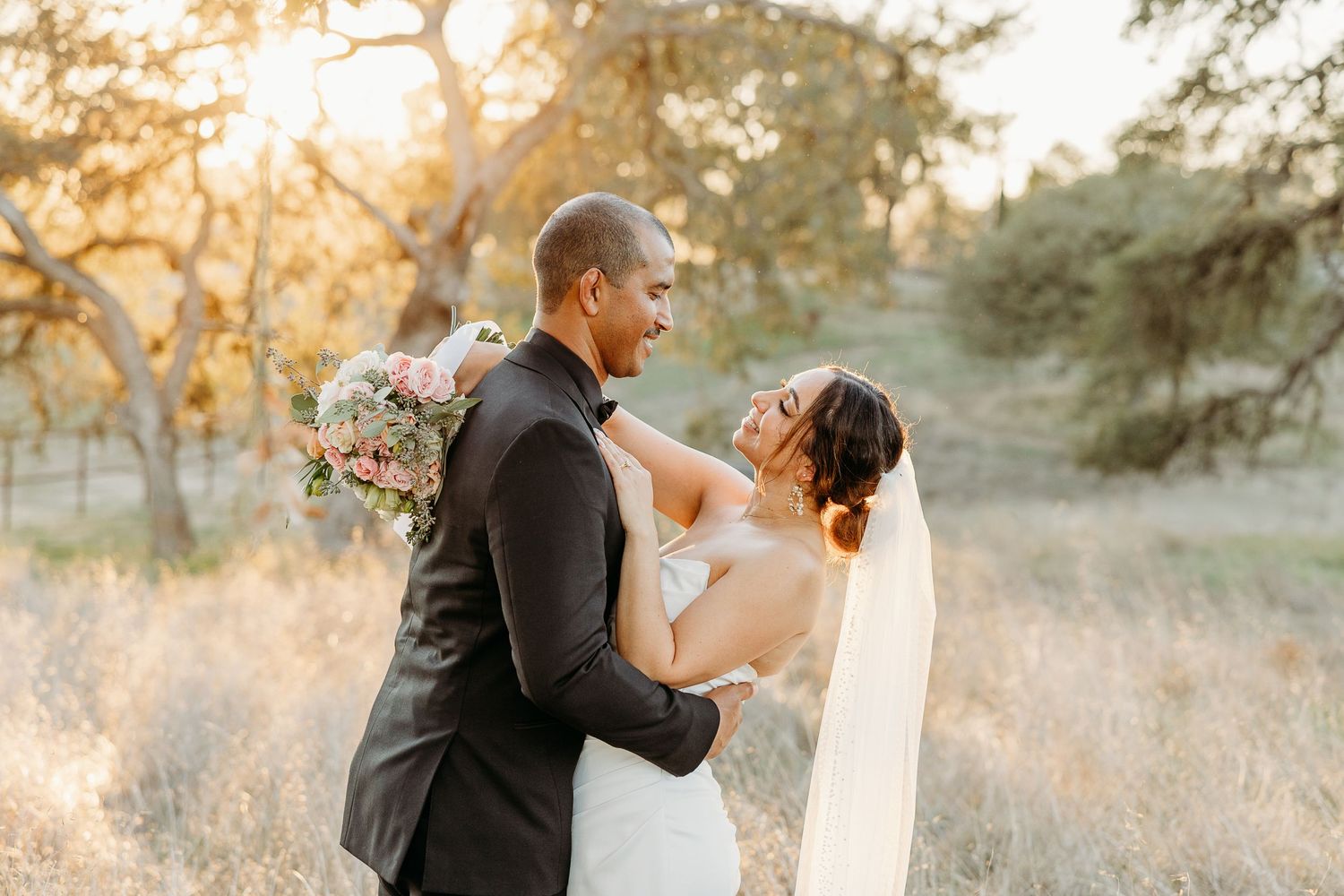 Romantic wedding couple embraces during golden hour sunset in a rustic outdoor field setting.