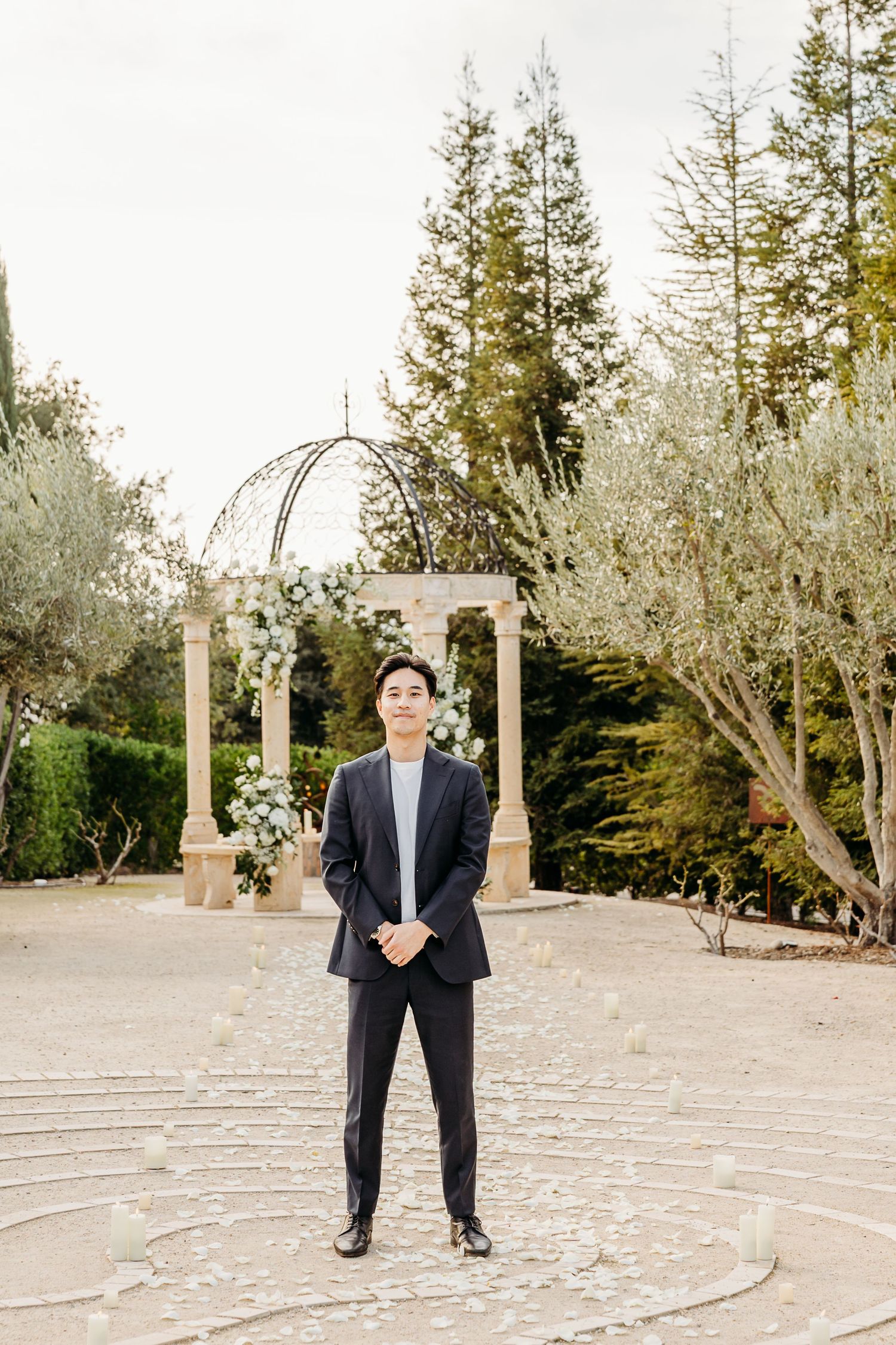 Stone labyrinth path surrounded by olive trees and columns in an outdoor garden setting.