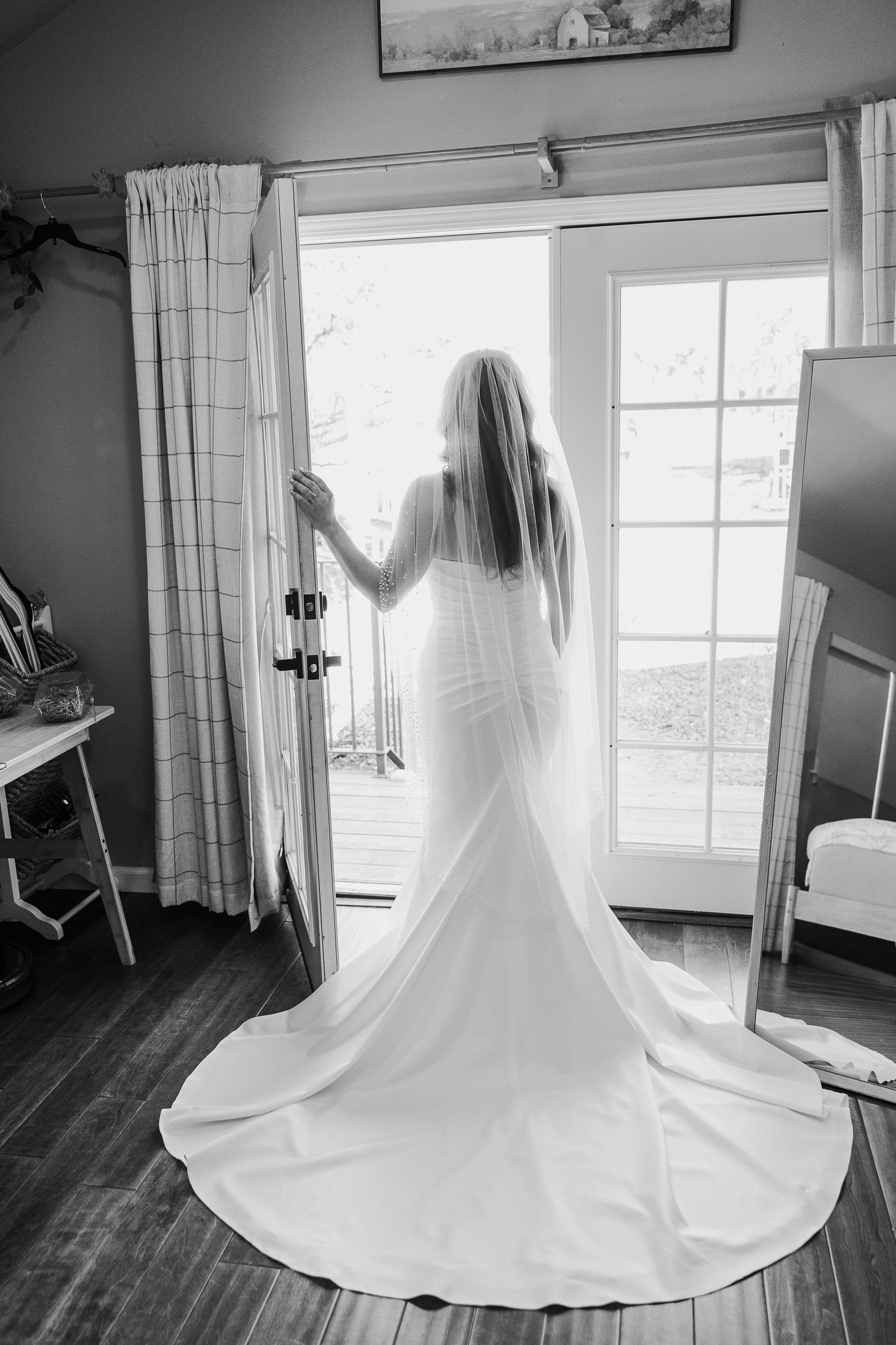 A flowing white wedding dress with a long train photographed in black and white against a window.