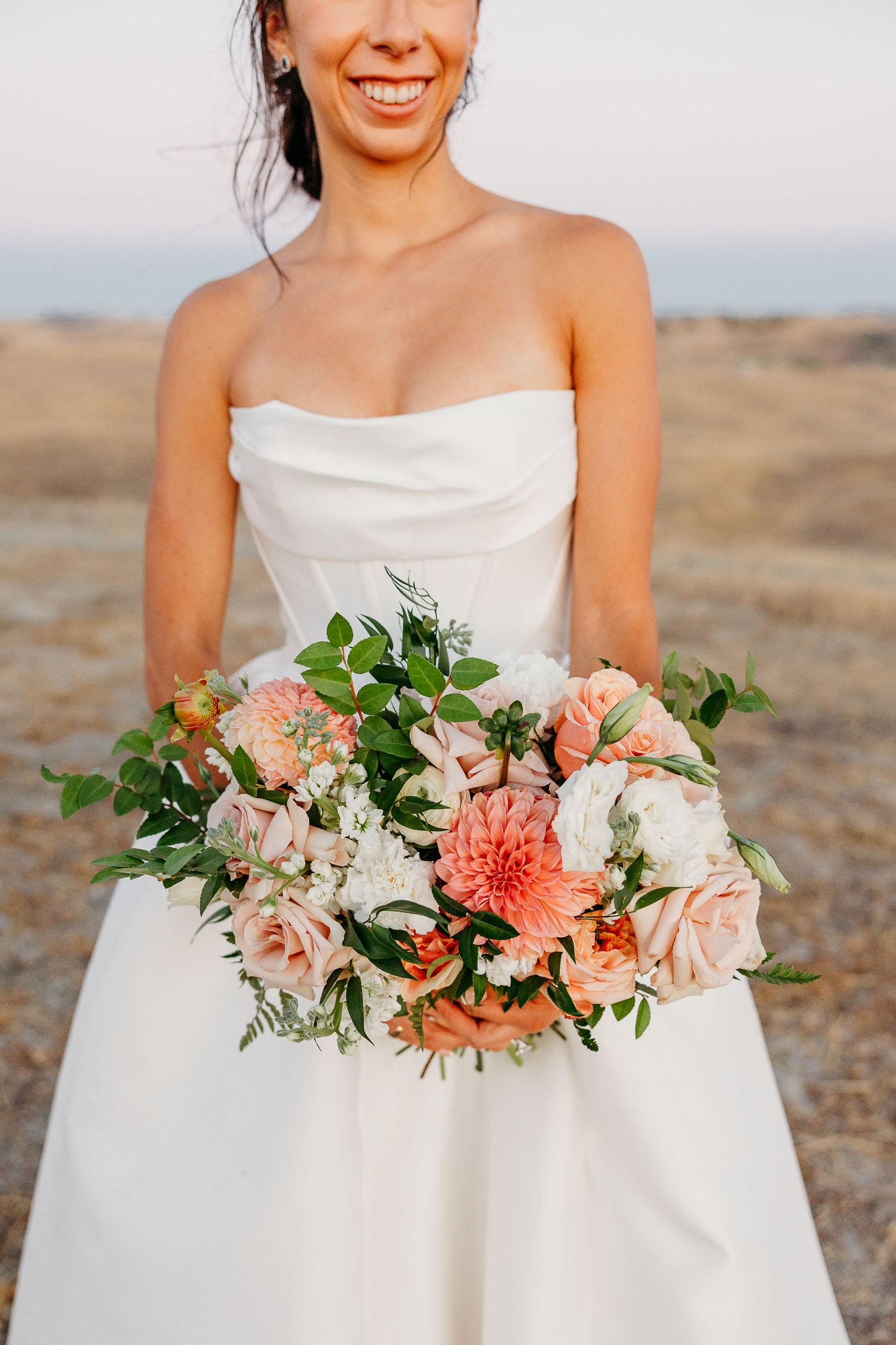 Beautiful coral and peach wedding bouquet with greenery against white wedding dress.