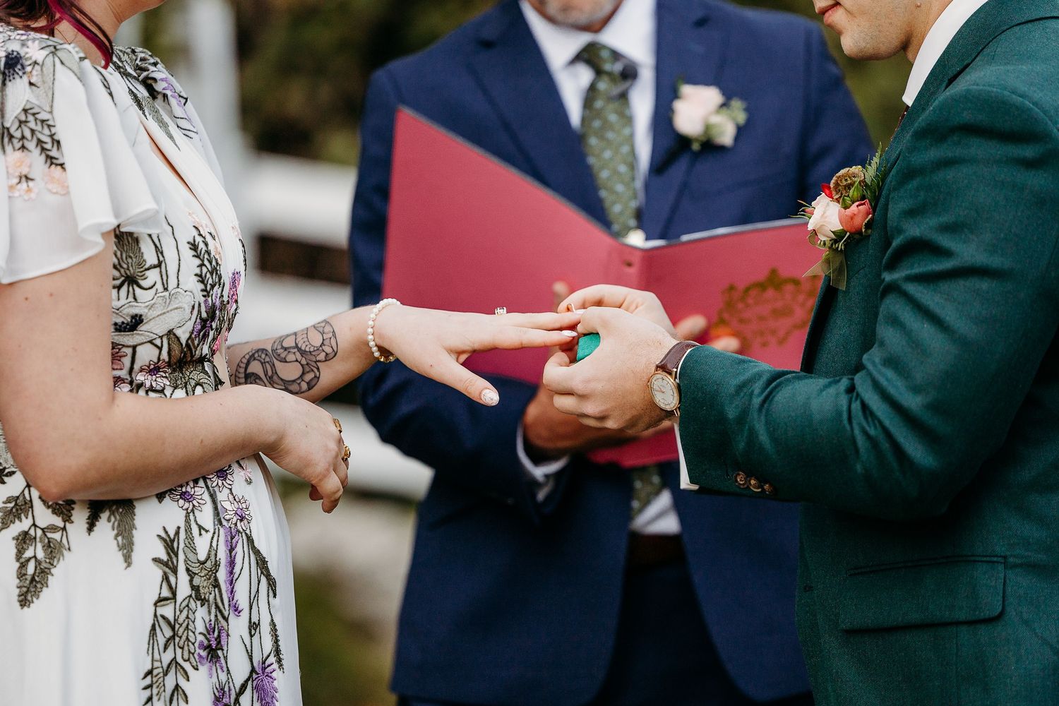 Wedding ceremony detail shot showing hands exchanging rings with boutonnieres visible.