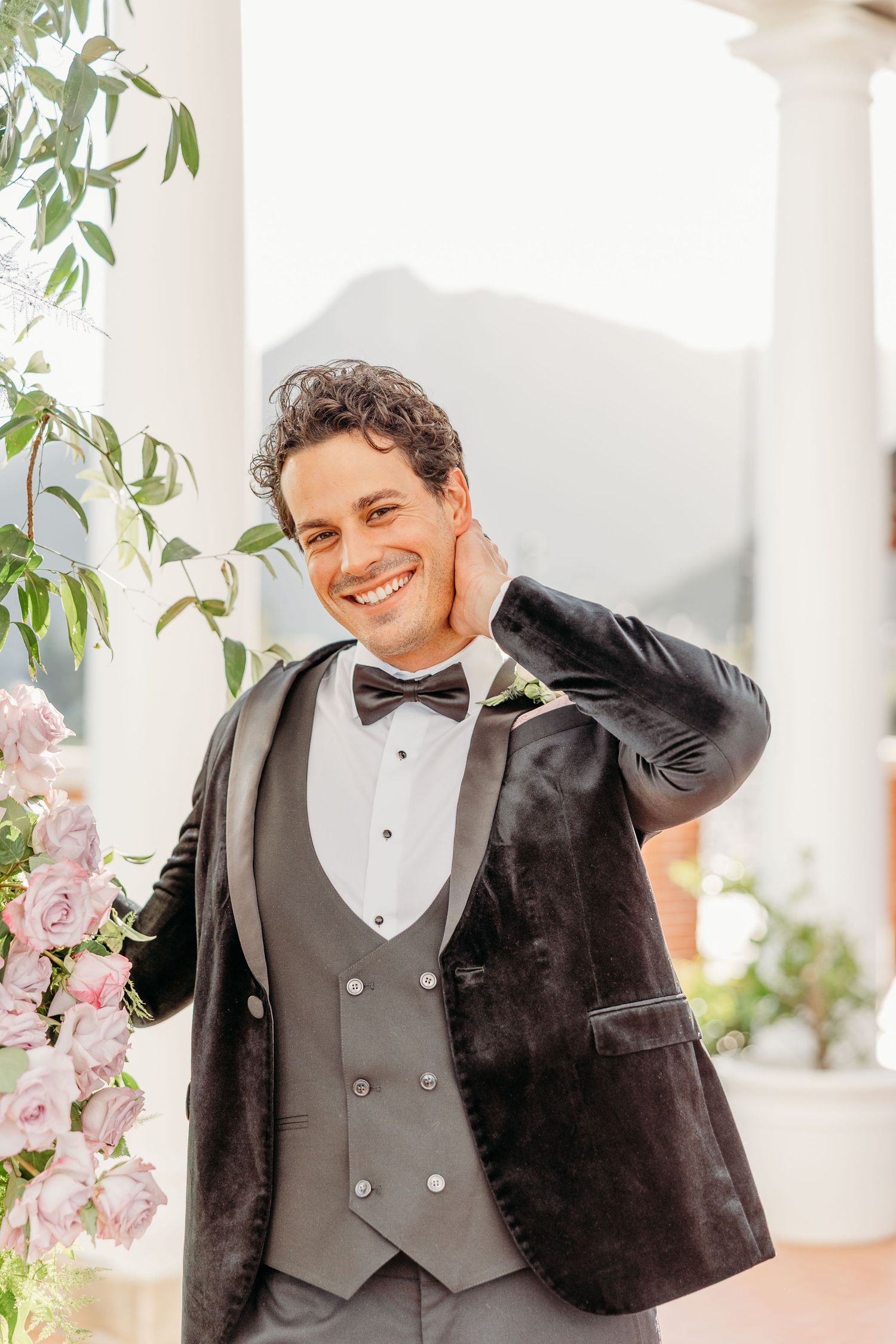 Groom in classic black tuxedo and bowtie smiles during outdoor wedding photoshoot.