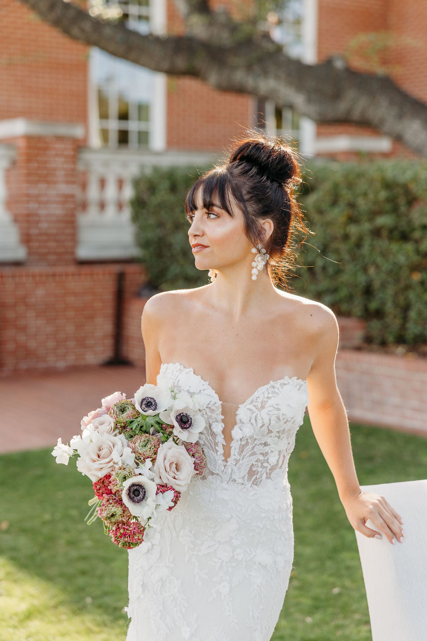 Bride in strapless lace wedding gown holds romantic bouquet of pink and burgundy flowers against brick building backdrop.
